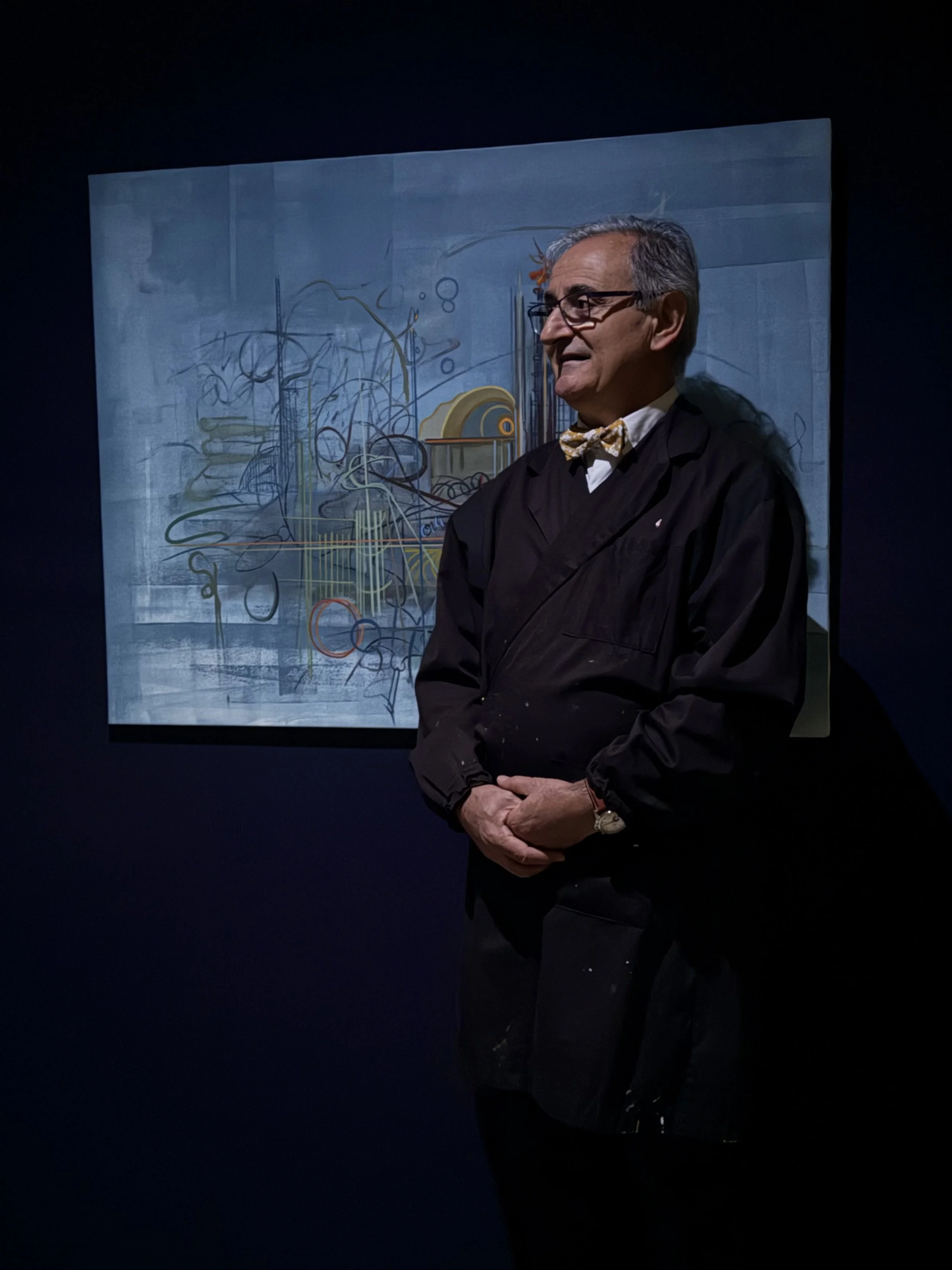 Jean-Pierre Lebel, un homme portant un costume noir, des lunettes et un nœud papillon, debout devant une peinture abstraite dans une galerie d'art.