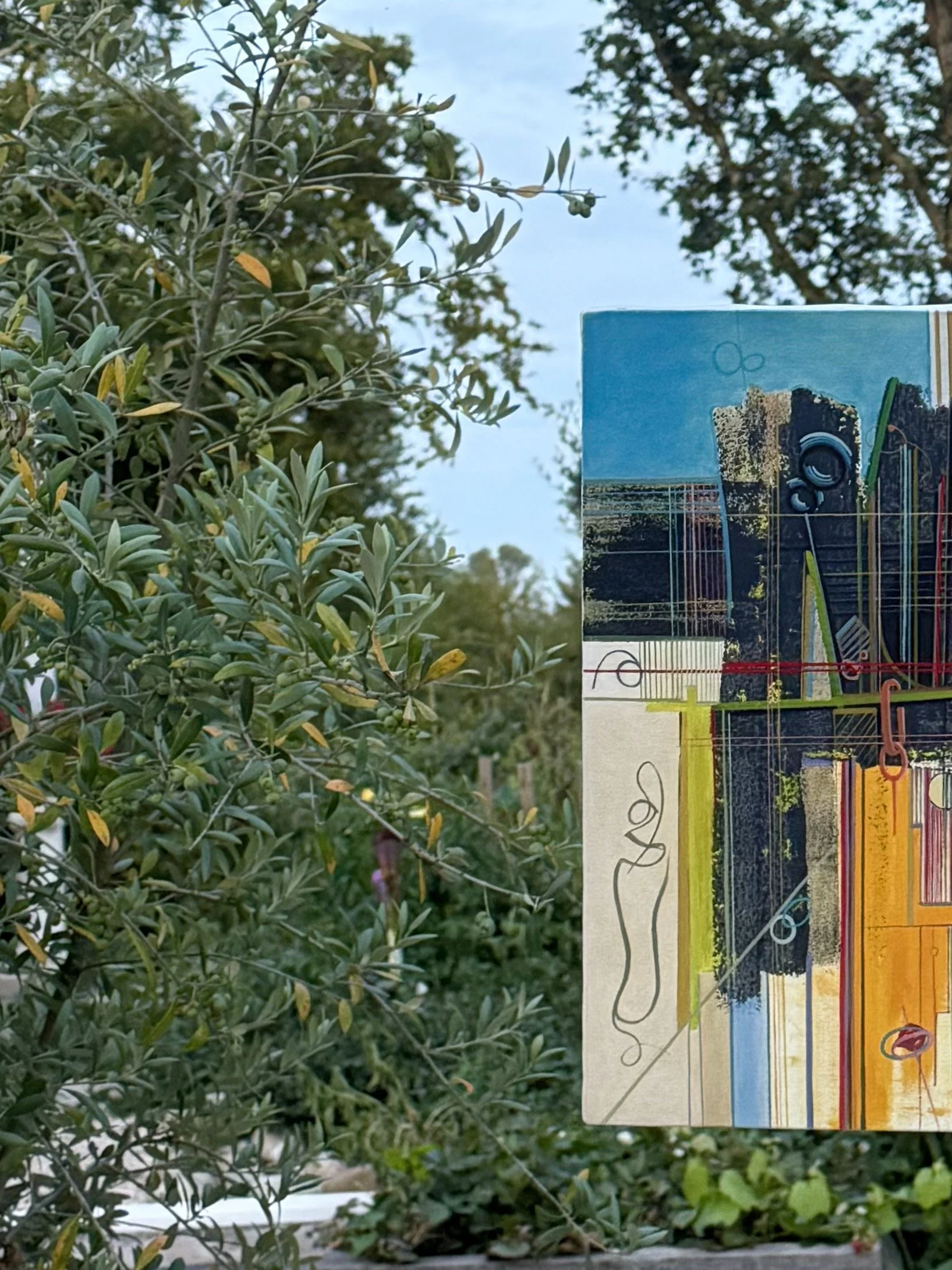 Un tableau coloré figurant des lignes abstraites, des formes géométriques, et des éléments de paysage avec du ciel bleu en arrière-plan, accrochée à une branche d'un arbre dans un jardin.