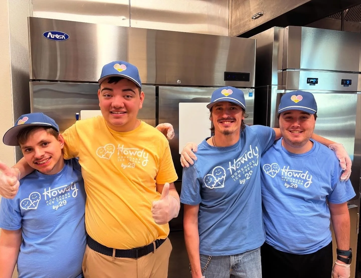 Four men wearing blue and yellow Howe Ice Cream shirts and matching hats, standing in front of industrial ice cream freezers, smiling and posing together inside an ice cream shop.
