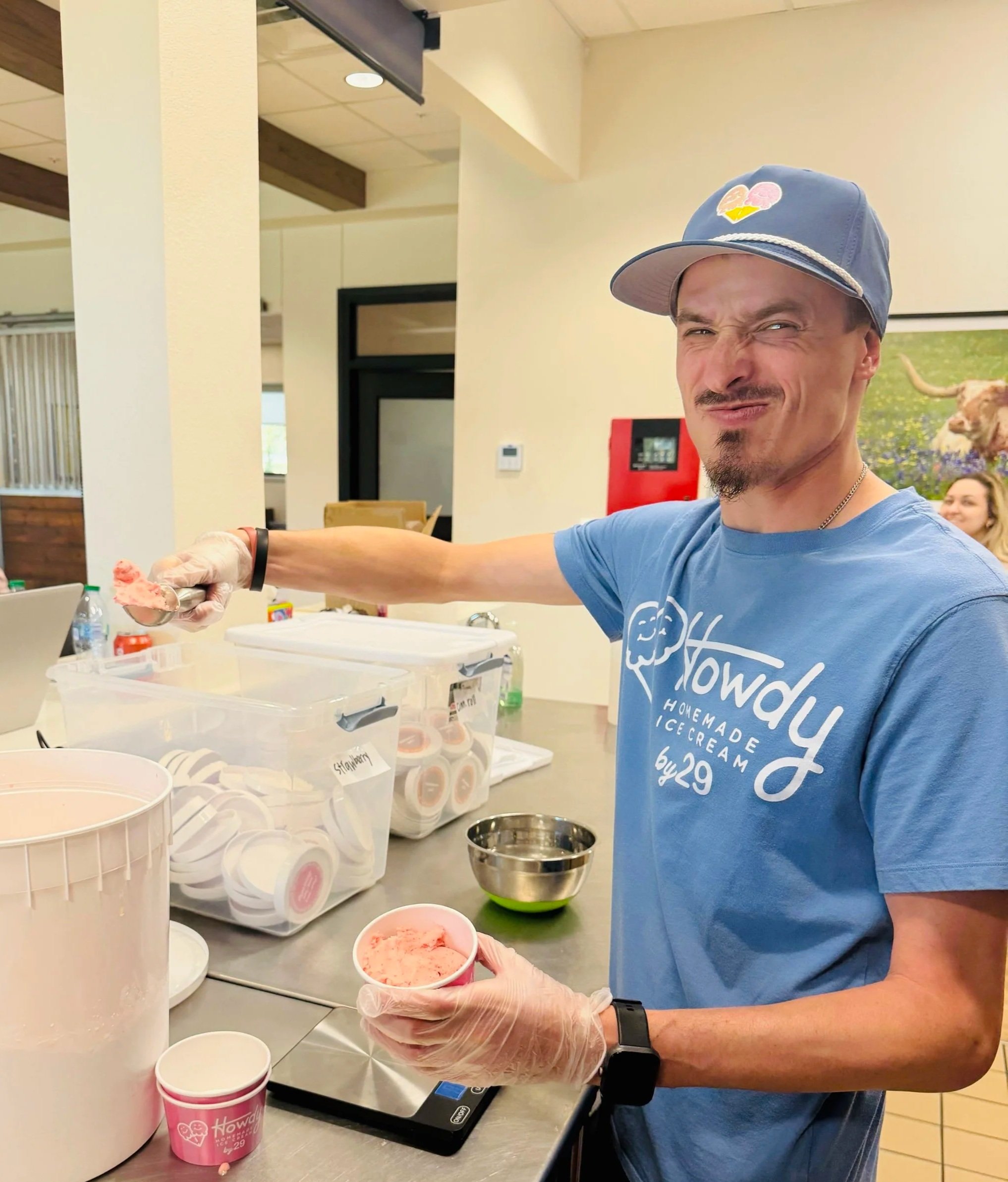 A man wearing a blue cap, blue t-shirt, and gloves is scooping ice cream into a pink cup at an ice cream shop. There are containers of ice cream, a metal mixing bowl, and various supplies on the counter around him. The background shows a cream-colored wall, a digital scale, and seating area.