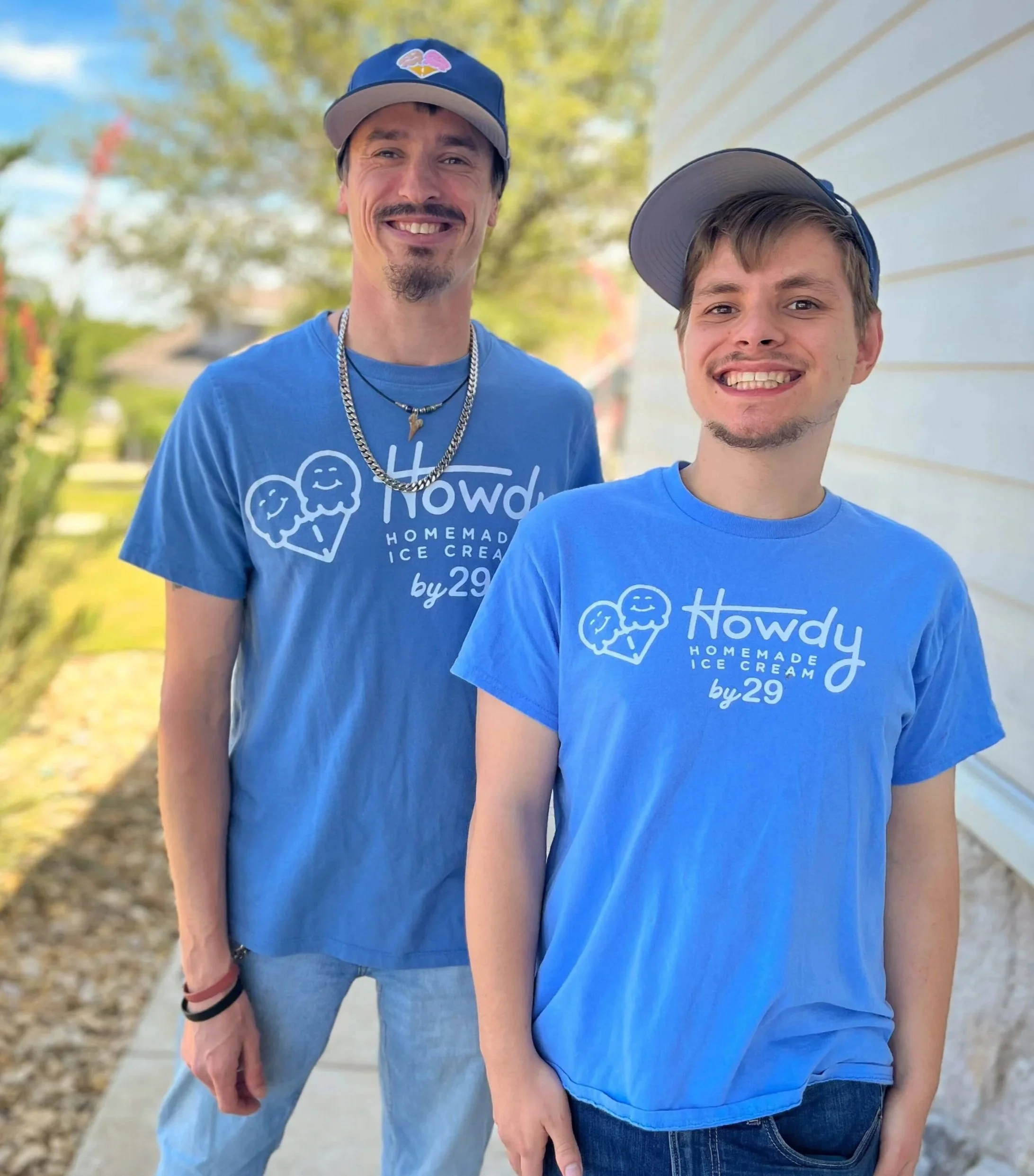 Two young men standing outdoors on a sunny day, smiling at the camera. Both are wearing blue t-shirts with a logo and text, and gray and blue caps. They are standing in front of a blurred background with trees and a white building.