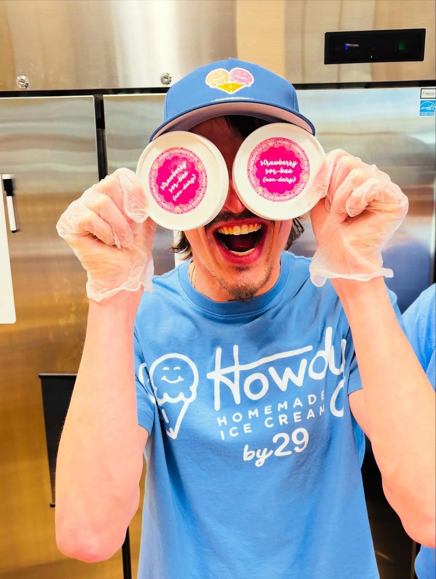 Person wearing a blue hat and gloves, holding two containers of strawberry ice cream in front of their face, in a commercial kitchen setting.
