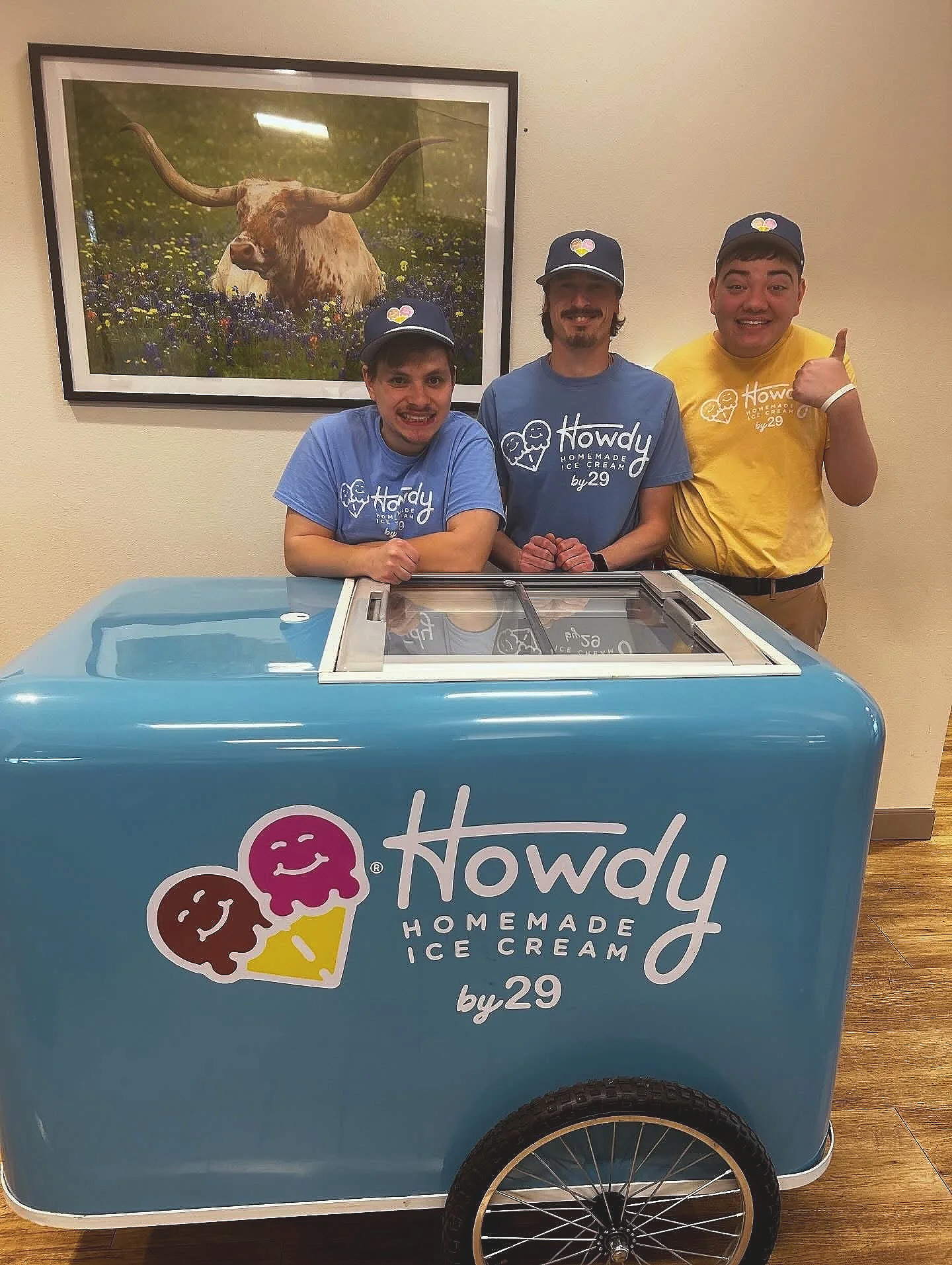 Three men standing behind a blue ice cream cart with the Howdy Homemade Ice Cream logo in front, smiling and wearing hats. They are inside a building, with a large framed picture of a longhorn cattle in a field hanging on the beige wall behind them.