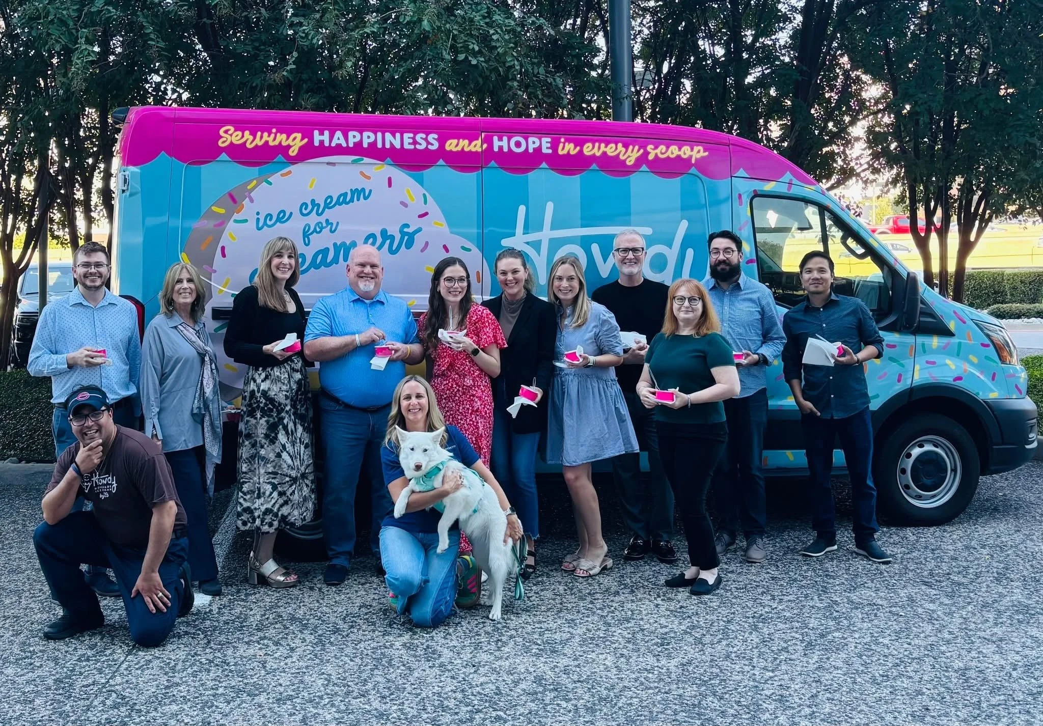 A group of people standing in front of a colorful ice cream truck during daytime. They are smiling and holding bowls of ice cream, with a dog sitting in front of the group. There are trees and parked cars in the background.