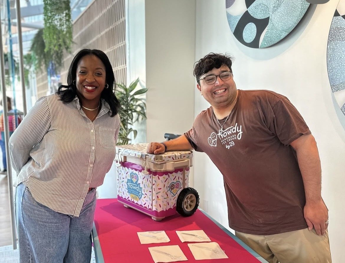 A woman and a man are standing indoors, smiling at the camera. They are posing next to a colorful ice cream cooler on a red table, with four paper bags in front of it. The woman is wearing a striped shirt and the man is wearing a brown t-shirt with a logo. The background shows a large window with greenery outside and decorative wall art.