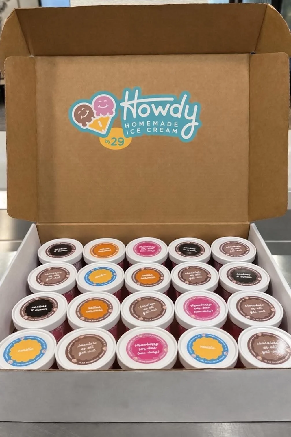 A cardboard box containing multiple small containers of homemade ice cream with colorful labels. The box has a sticker on the inside of the lid that reads '.Howdy Homemade Ice Cream by 29' and features two cartoon ice cream cones with smiling faces.