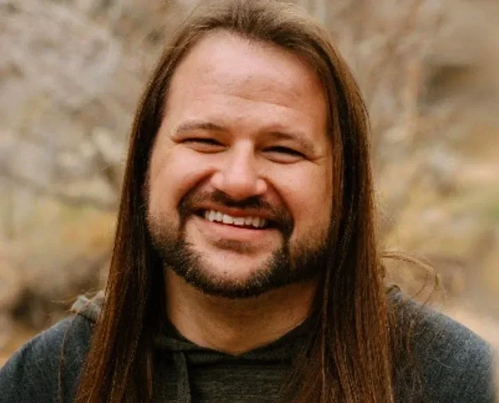 Therapist. A smiling man with long brown hair and a beard outdoors.