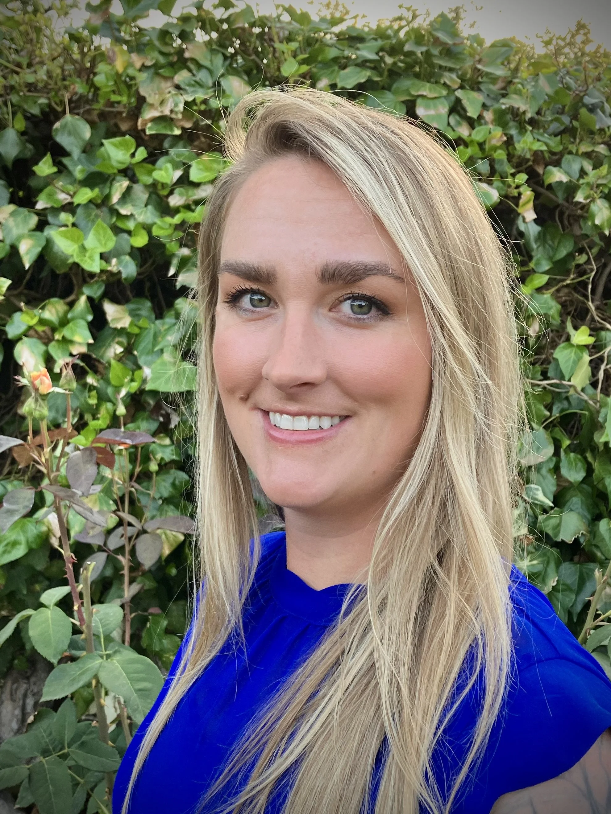 A young woman with long blonde hair, wearing a blue top, smiling outdoors in front of green leafy bushes.