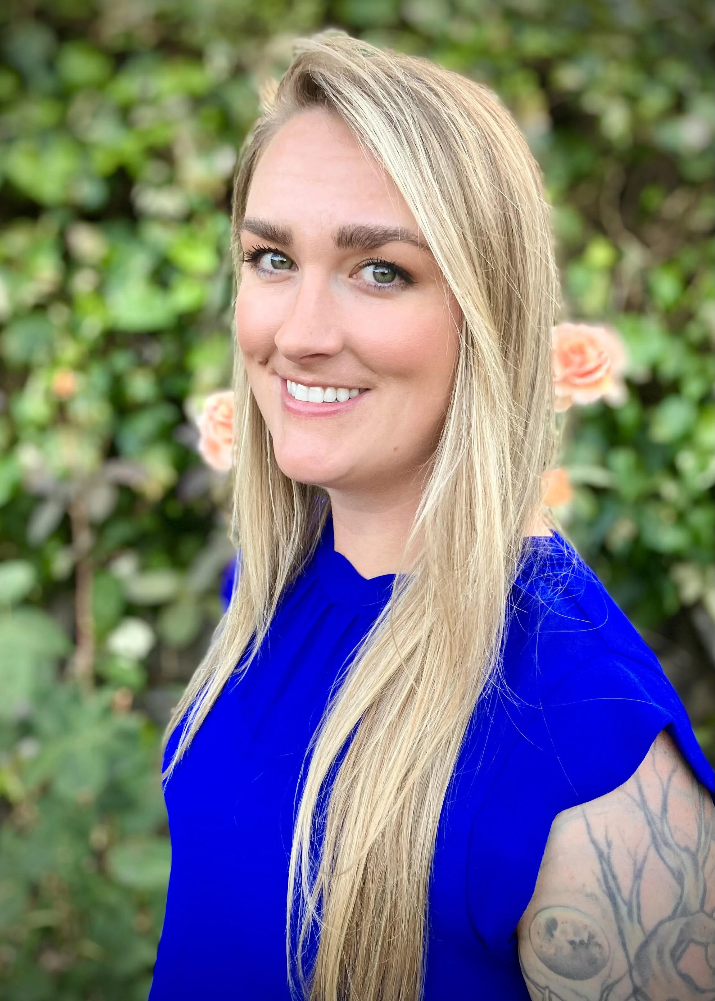 A woman with long blonde hair, green eyes, and a bright smile, wearing a royal blue top, standing outdoors with a background of green foliage and pink roses.