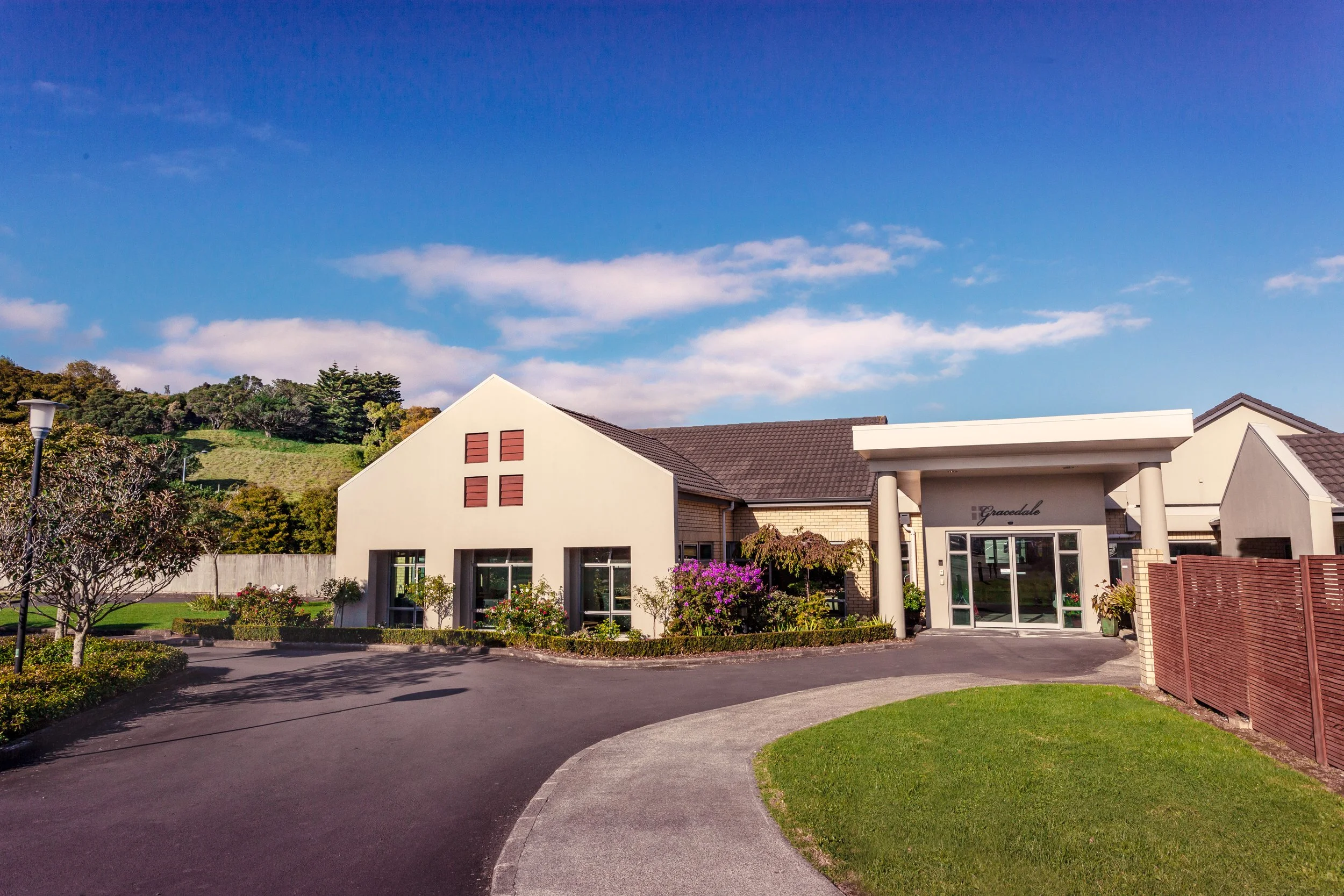 A modern aged care facility with a landscaped front yard, trees, and a driveway, under a partly cloudy blue sky.