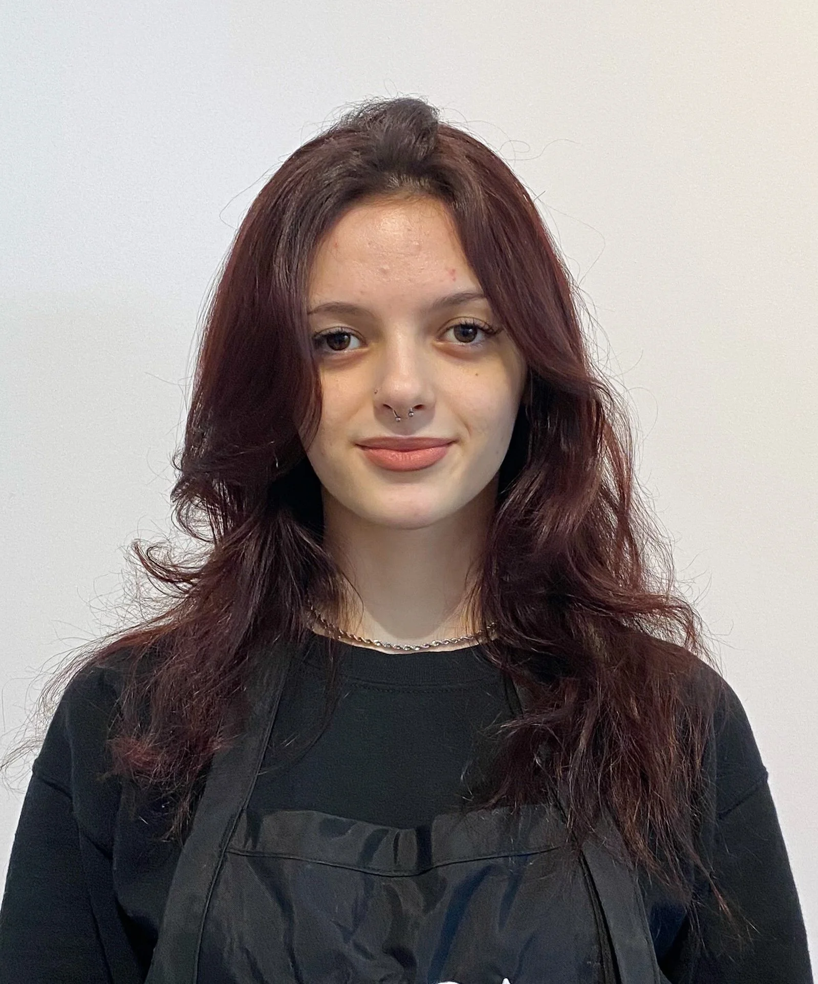 Portrait of a young woman with wavy, auburn hair, light skin, and a septum piercing, wearing a black shirt and a chain necklace, standing against a plain white background.