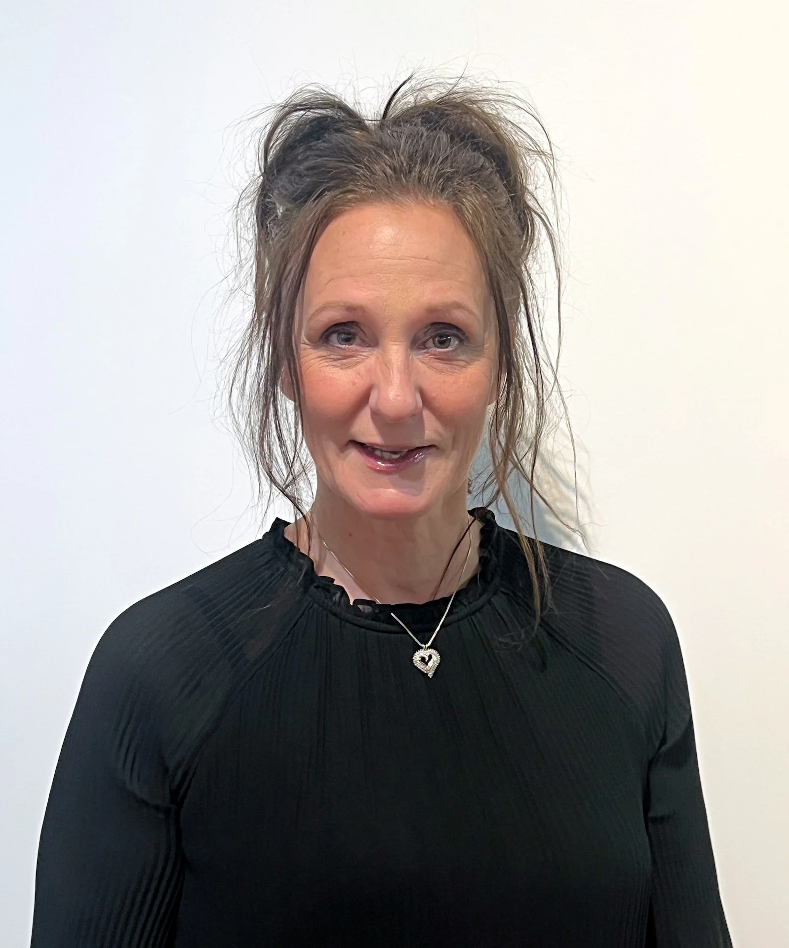 A woman with curly, messy hair in a bun, wearing a black top and a silver heart-shaped necklace, standing against a plain white background.