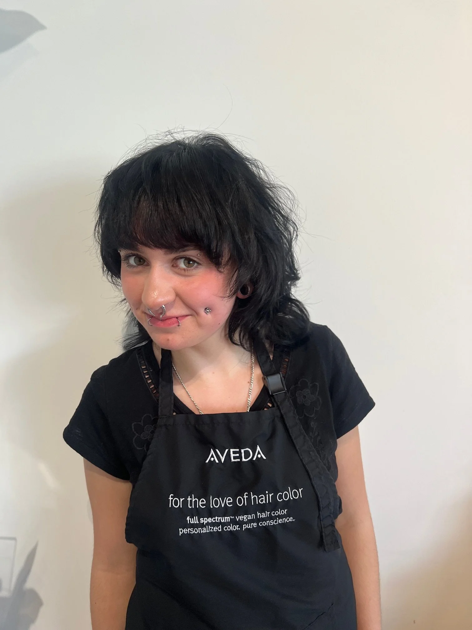 Woman with dark, wavy hair and multiple facial piercings, wearing a black apron with 'Aveda' text, standing against a plain wall.