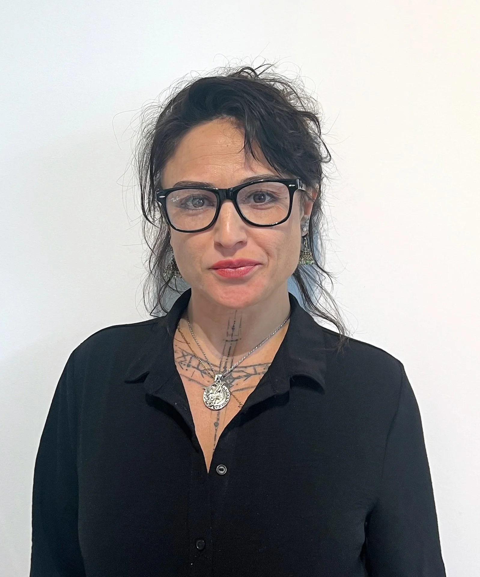 A woman with wavy dark hair and glasses, wearing a black shirt, silver jewelry, and earrings, standing against a white background.