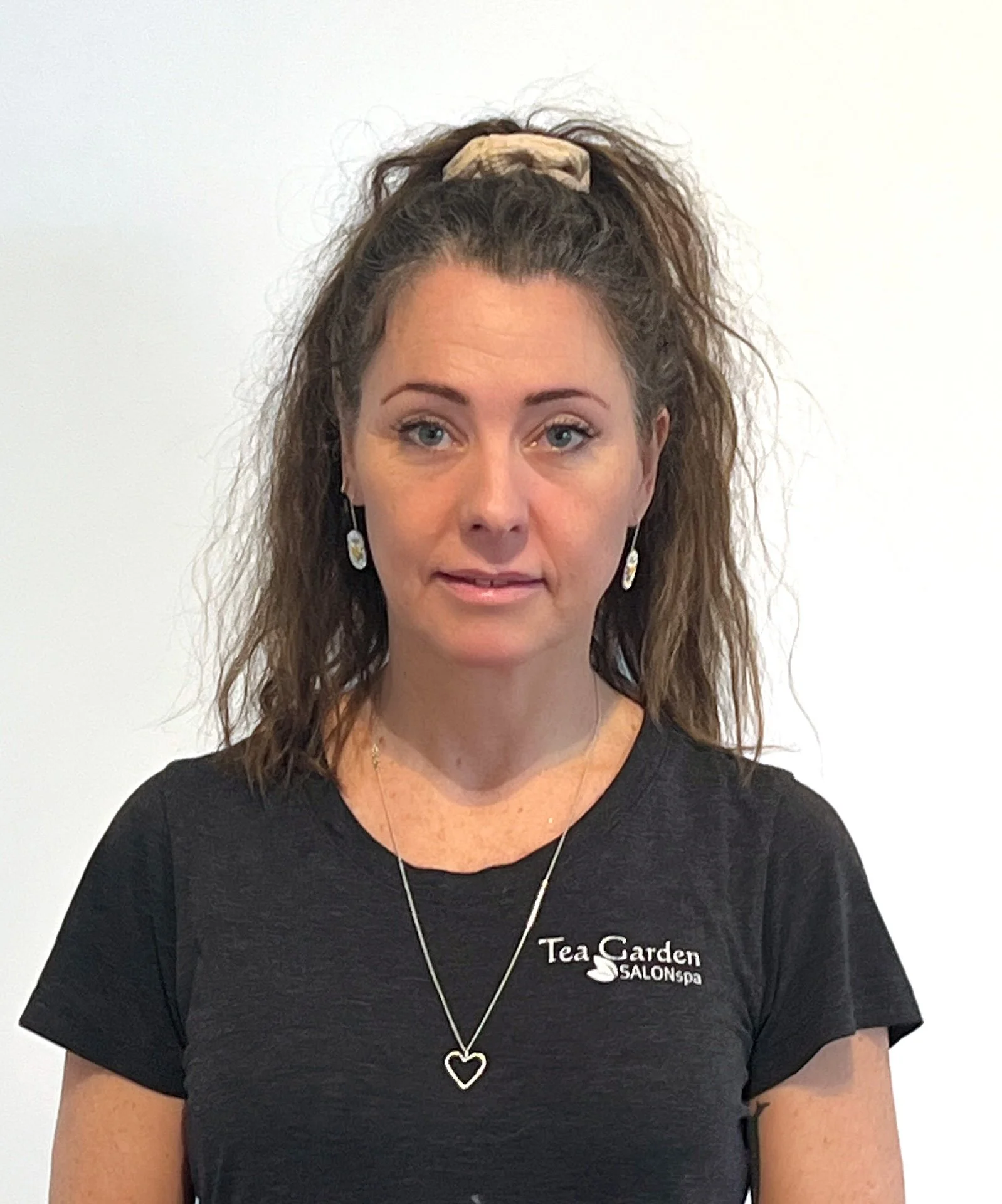 Woman with wavy brown hair tied in a high ponytail, wearing earrings and a black t-shirt with 'Tea Garden SALONspa' logo, against a plain white background.