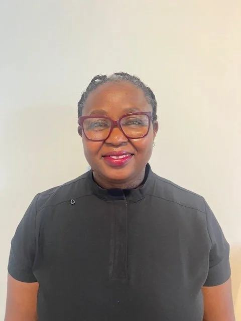 A woman with dark skin, wearing red glasses, smiling, and dressed in a black top with a high collar, posing against a plain, light-colored background.