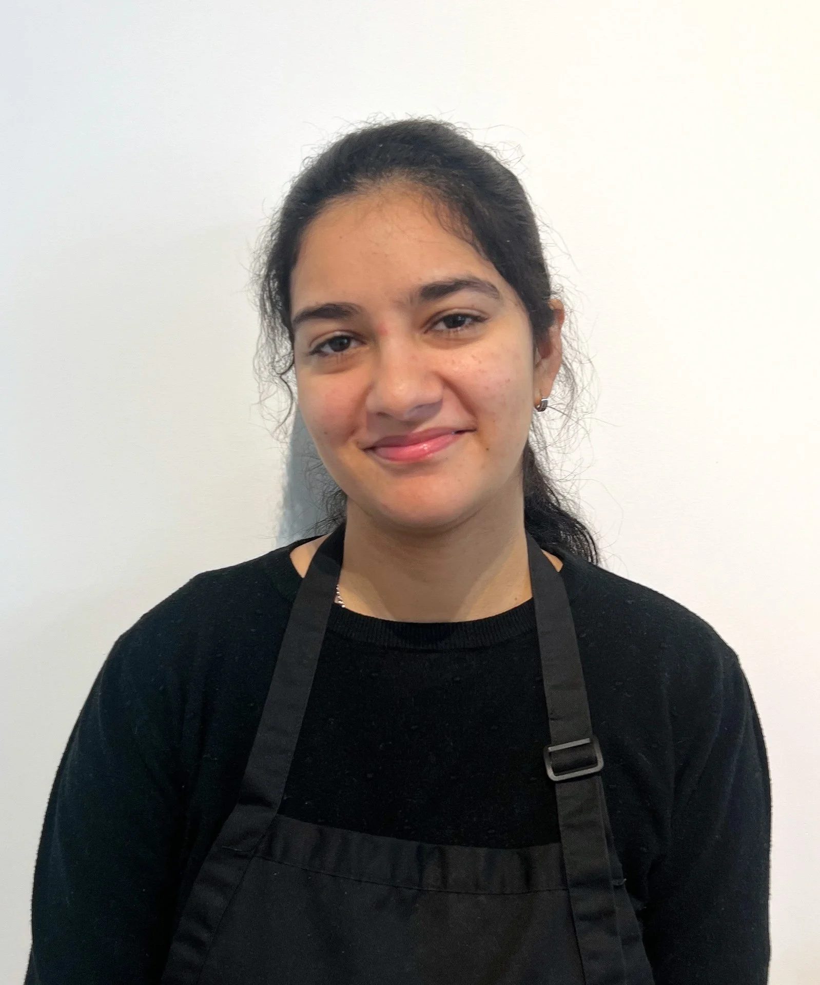 A young woman with dark hair, wearing a black shirt and apron, smiling against a plain white wall.