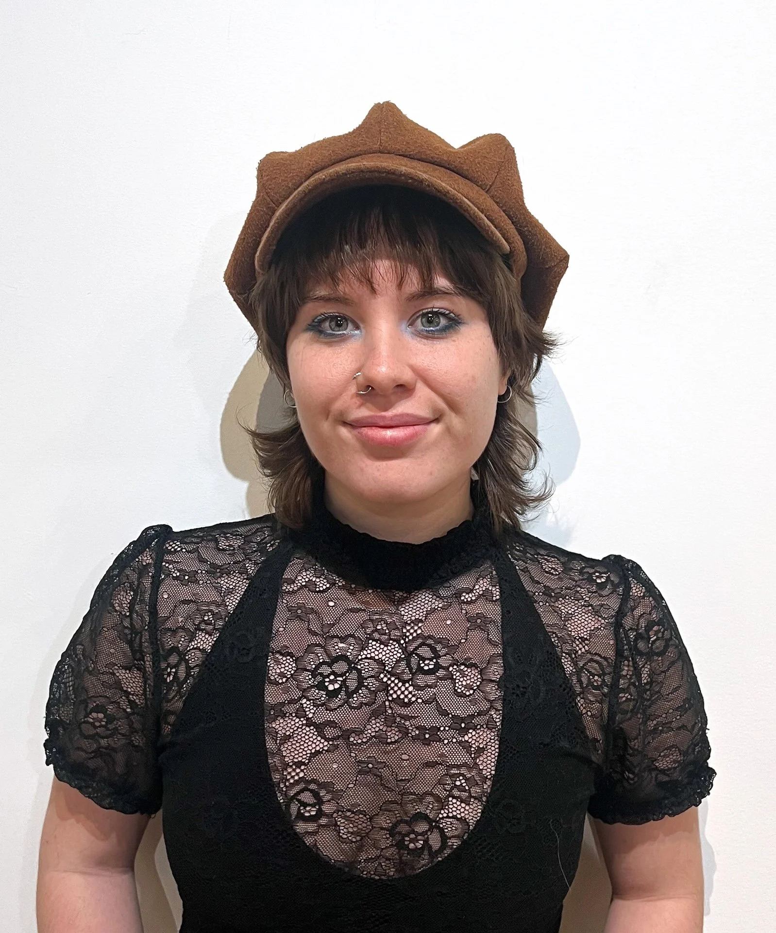 Young woman wearing a brown hat and black lace top, standing against a white background.