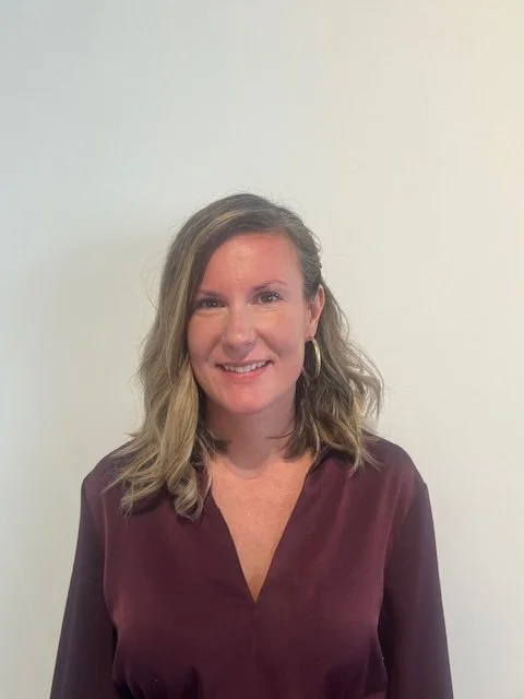 A woman with shoulder-length wavy blonde hair, wearing a maroon blouse with a V-neck and hoop earrings, smiling at the camera against a plain white background.