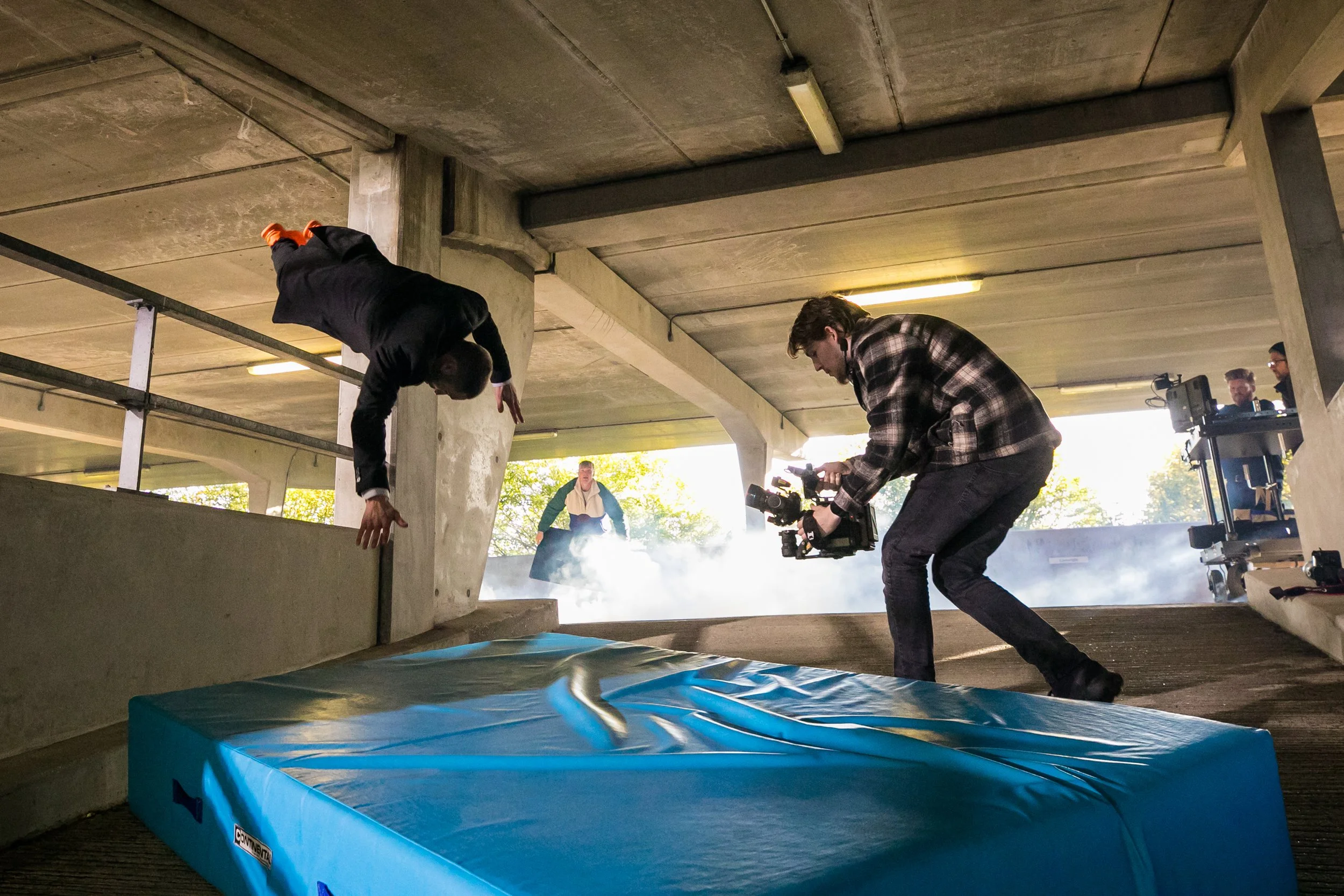 Filming crew capturing a stunt scene with a man falling onto a crash pad in an underpass parking garage.