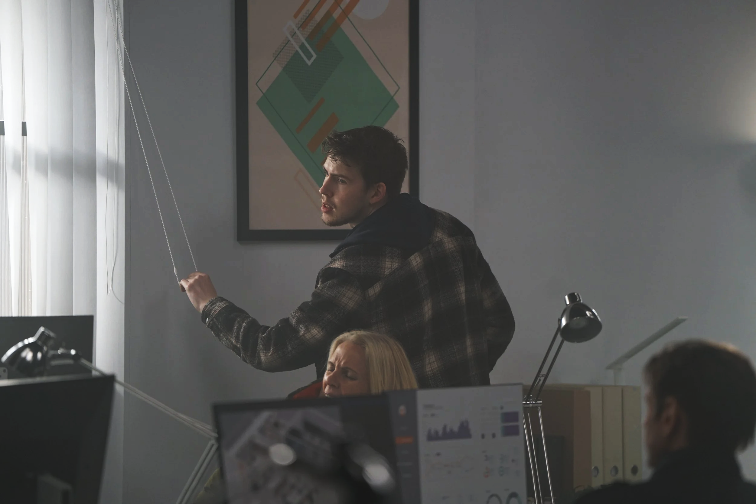 A man standing in an office, pointing at blinds. Two women and another man are seated at desks working on computers.
