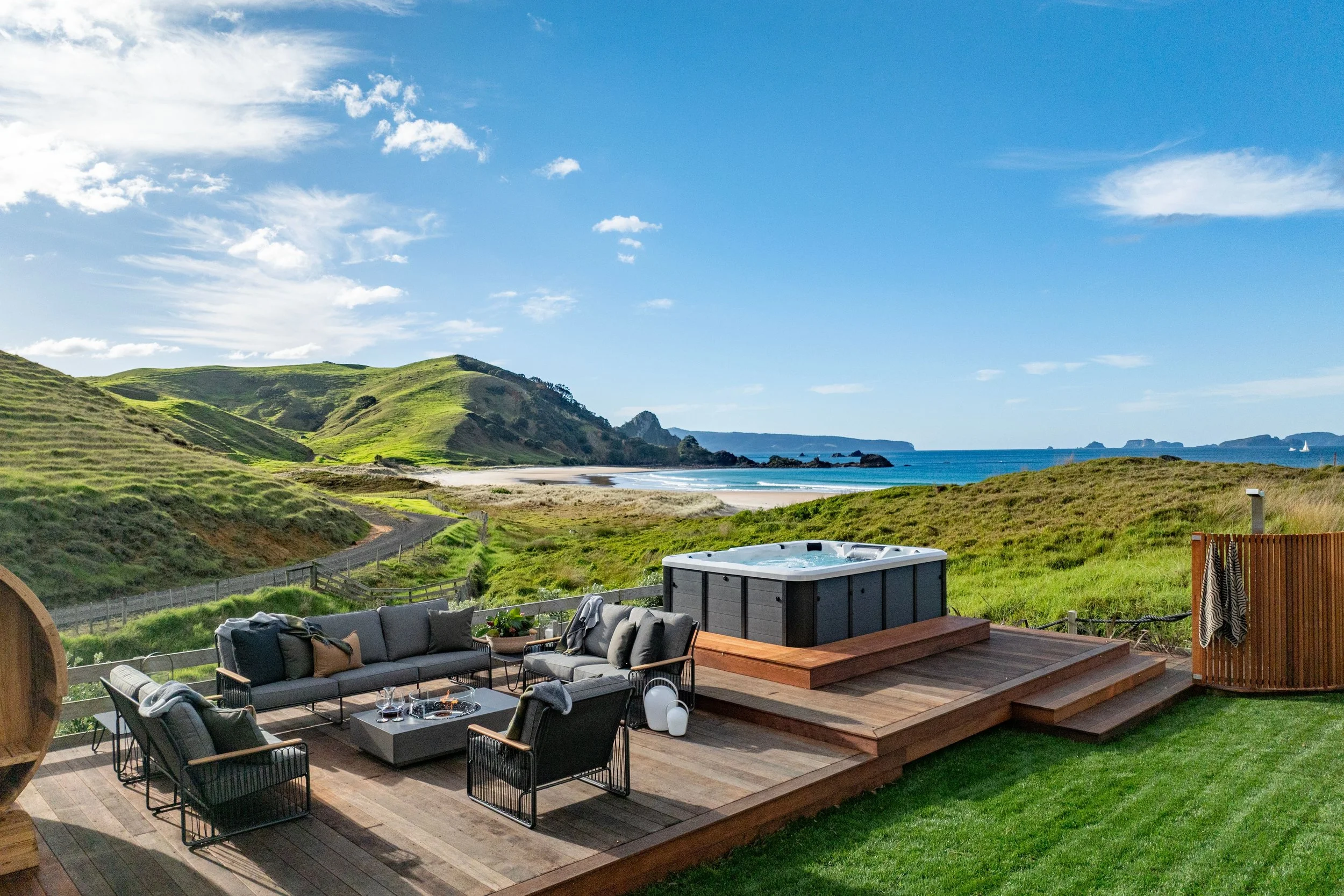Outdoor deck, sauna, seating area with a fire pit, hot tub, and outdoor shower, overlooking green hills at Opito Point Lodge - Luxury Accommodation in Coromandel, New Zealand