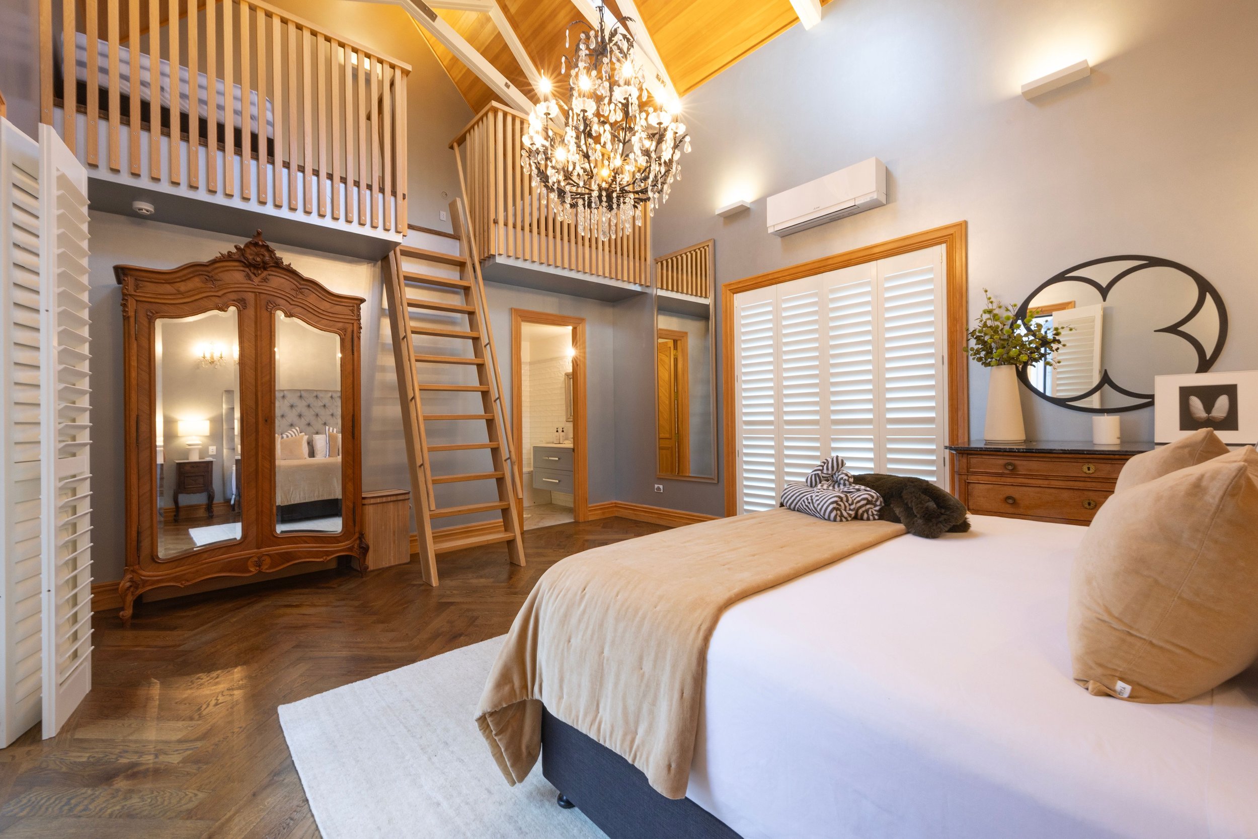 Luxurious bedroom with high ceiling, chandelier, wooden loft, and elegant furniture, including a large bed with beige bedding, wooden dresser, and ornate wardrobe, decorated with modern art and lush plants.
