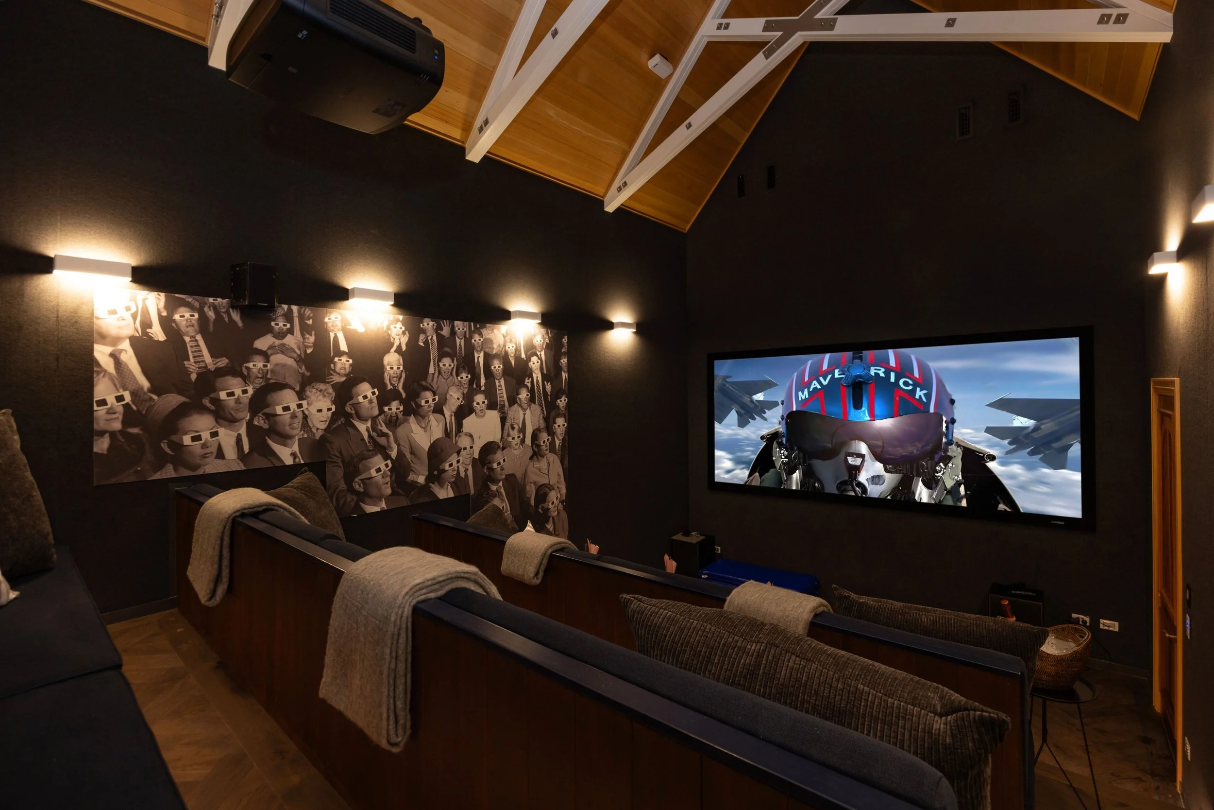 Home theater room with black walls, featuring a large slide screen showing a jet fighter cockpit view, and a black-and-white photo mural of people watching a 3D movie, all illuminated by wall-mounted lights at Opito Point Lodge, luxury beachfront acc