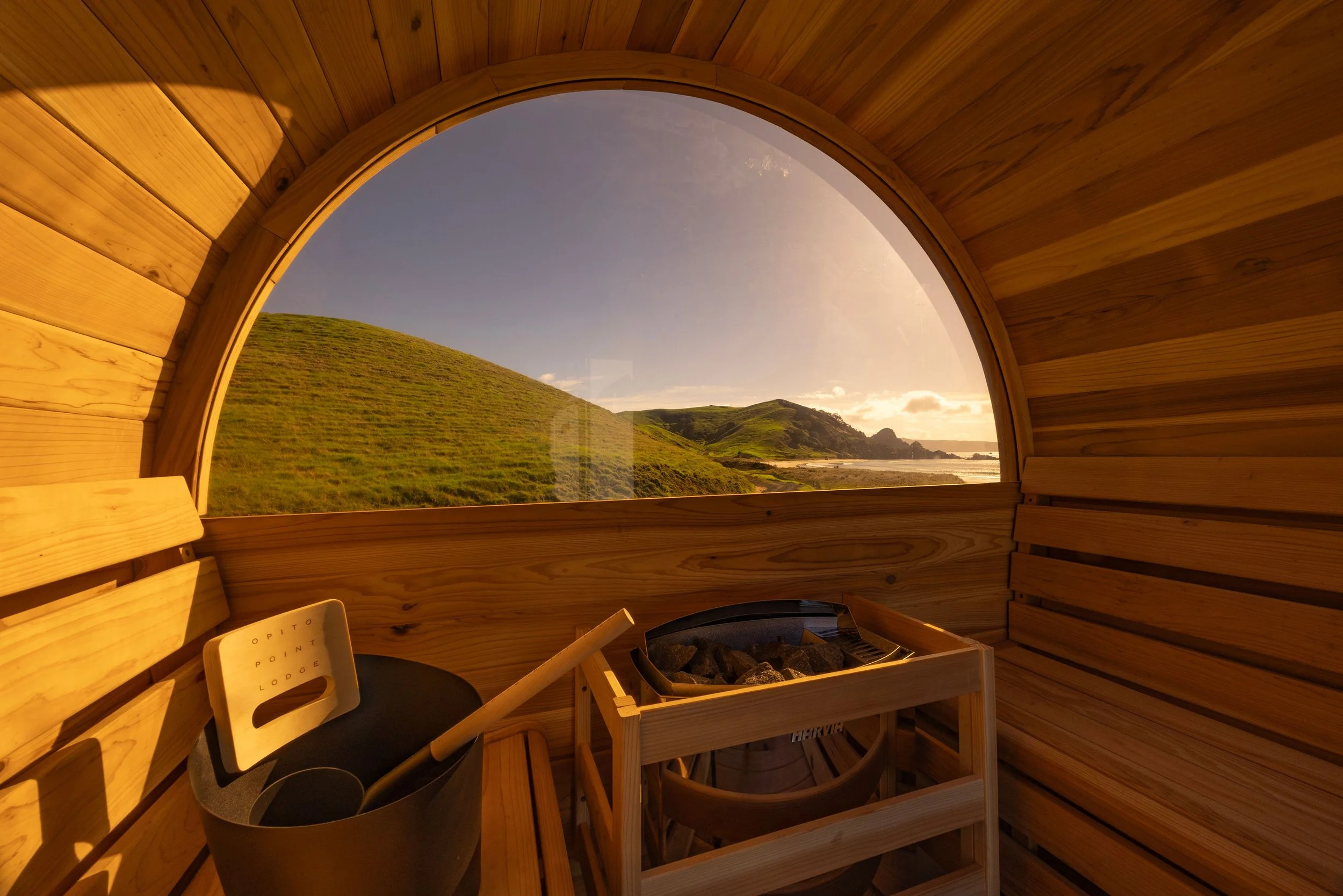Interior of a wooden sauna with a large window showing a scenic view of green hills, a coastline, and the ocean during sunset at Opito Point Lodge, luxury beachfront accommodation.