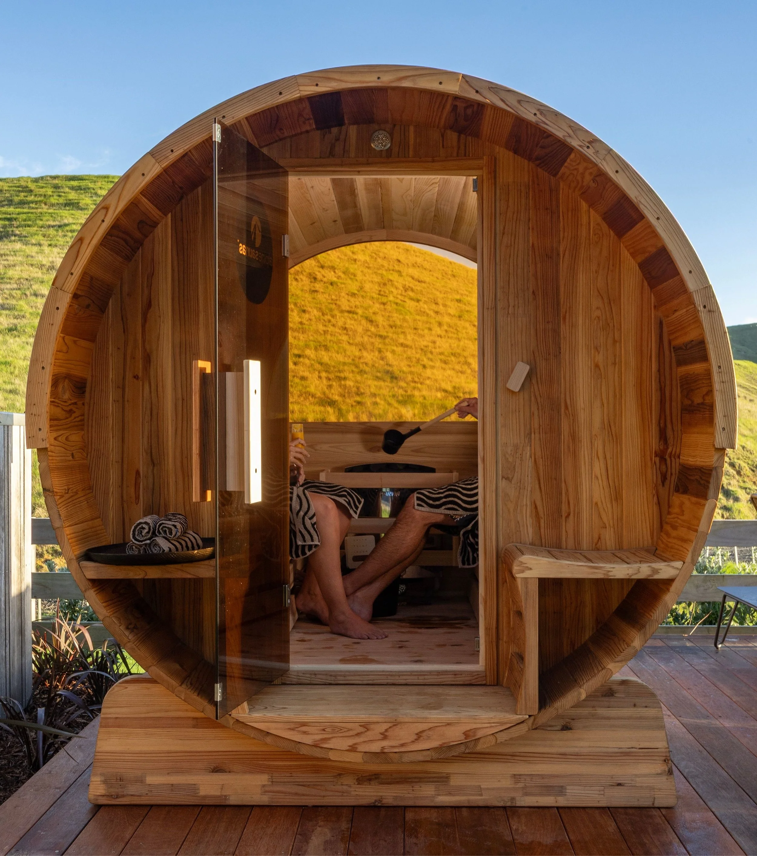 A wooden barrel-shaped sauna with a glass door, located outdoors on a deck with a scenic mountain background, and two people inside relaxing and enjoying the warm environment at Opito Point Lodge, luxury beachfront accommodation.