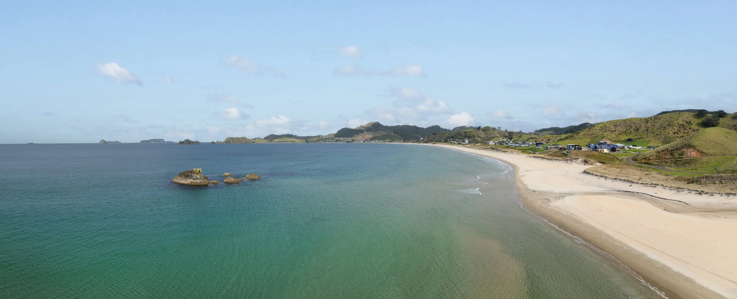 A scenic view of a sandy beach with clear greenish-blue water, rocky formations, and a small shoreline. There are rolling green hills with houses and a partly cloudy sky in the background. at Opito Point Lodge, luxury beachfront accommodation.