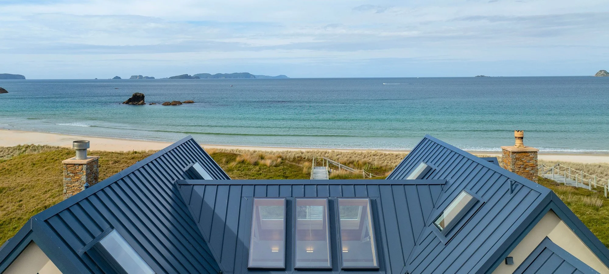 View of the ocean with the beach in the foreground and rooftops of a house with skylights in the bottom part of the image at Opito Point Lodge, luxury beachfront accommodation.