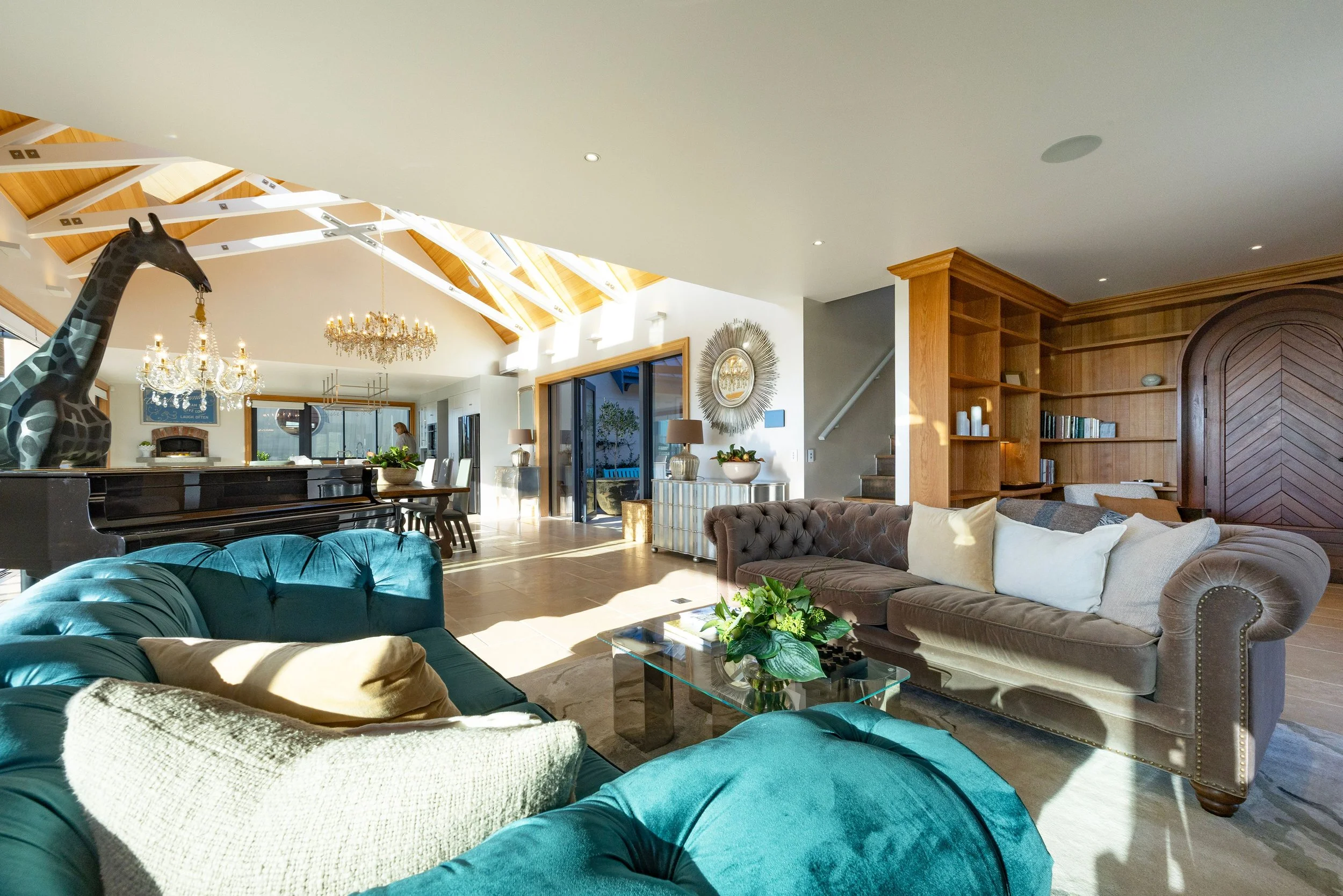 Spacious living room with high vaulted ceiling, large windows, and wooden accents. Feature dining area with a chandelier at Opito Point Lodge, luxury beachfront accommodation