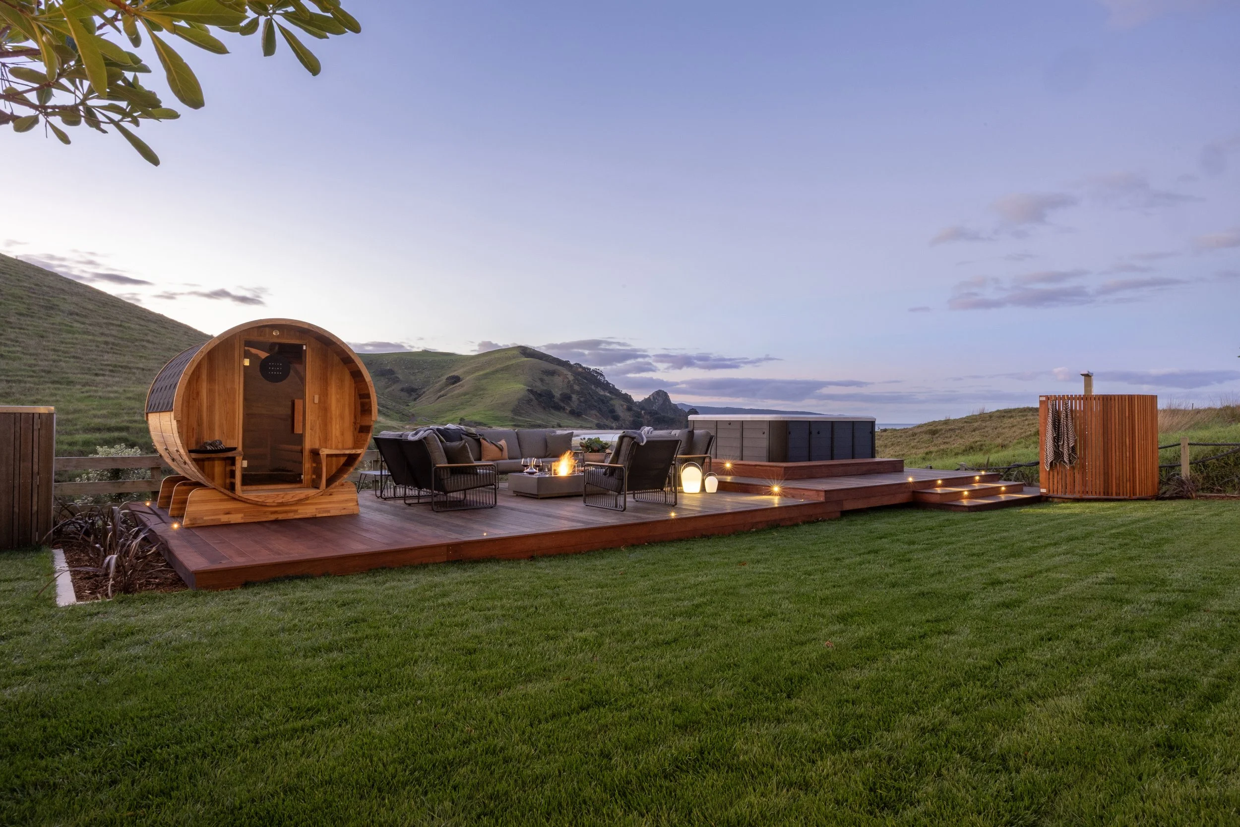 Outdoor deck with a sauna, outdoor seating area with a fire pit, hot tub, and outdoor shower, set against a backdrop of green rolling hills and a cloudy sky at dusk at Opito Point Lodge, luxury beachfront accommodation.