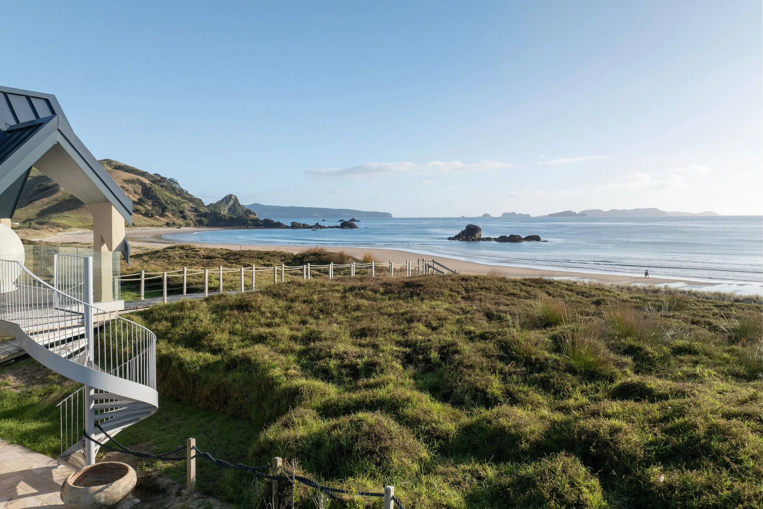 View of a sandy beach with gentle waves, rocks in the water, a grassy dunes and hills, and a clear blue sky, with part of a building and a spiral staircase in the foreground at Opito Point Lodge, luxury beachfront accommodation.