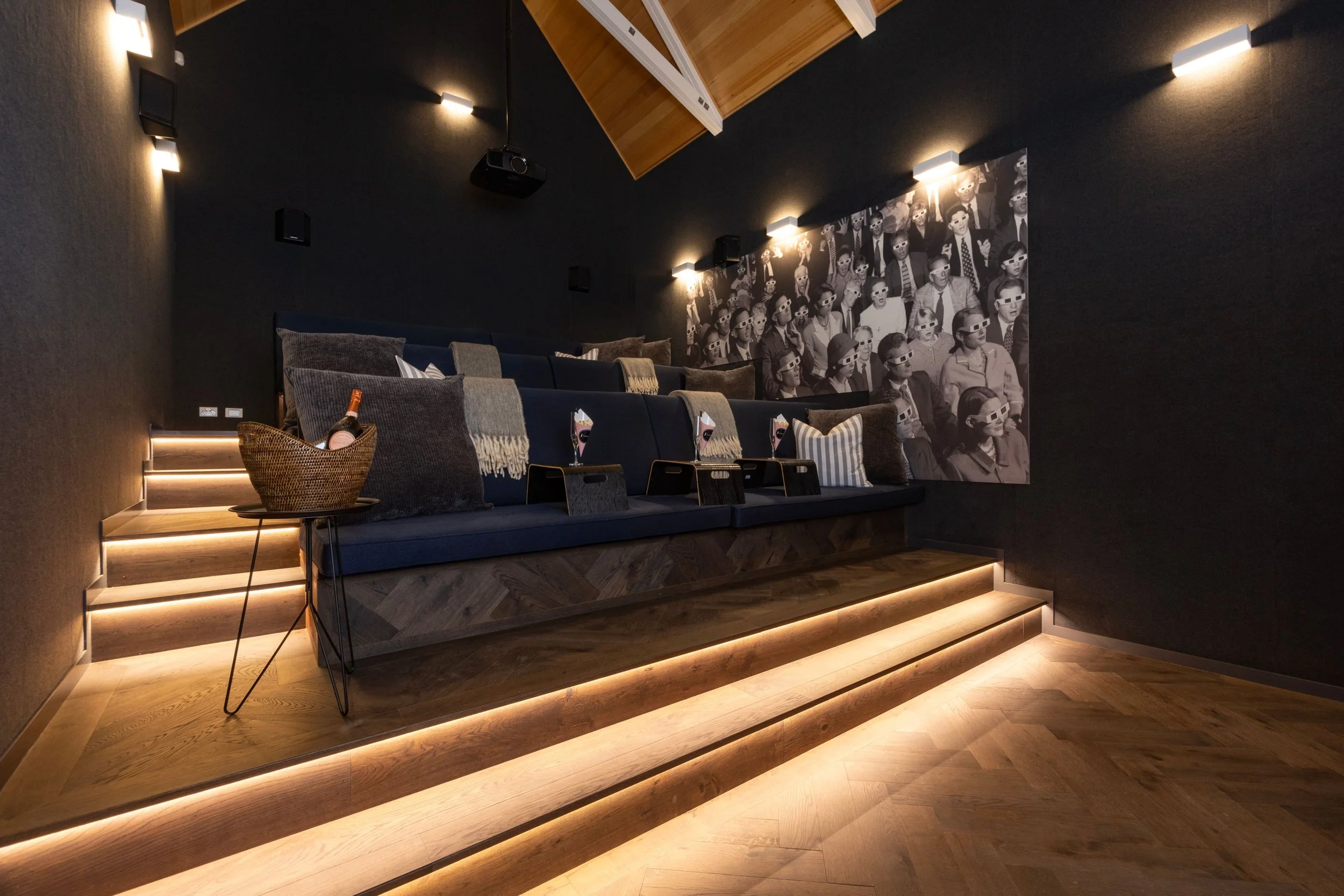A modern, dimly lit home theater with a black wall featuring a large black and white photo of people wearing 3D glasses. The theater has a tiered seating area with dark cushions and throw pillows, illuminated by built-in strip lighting along the stai