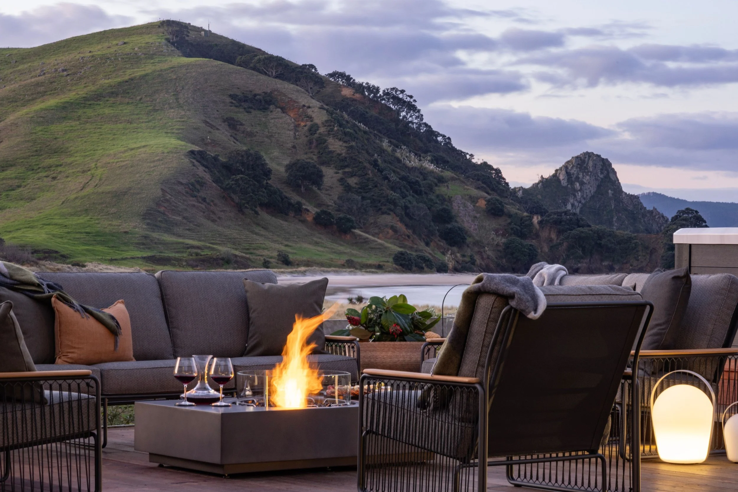Outdoor patio with seating area, a fire pit, and glasses of red wine, overlooking a mountain and a beach in the background at dusk at Opito Point Lodge, luxury beachfront accommodation.