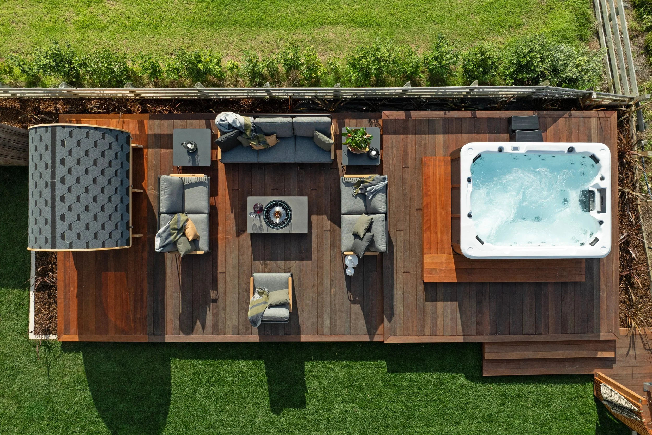 Aerial view of a wooden deck with outdoor furniture, a hot tub, and a small pergola, surrounded by grass and a fence at Opito Point Lodge, luxury beachfront accommodation.