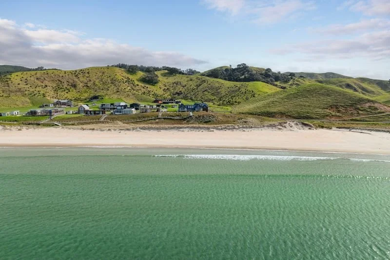 A beach with green water, a sandy shoreline, and green hills with houses in the background under a partly cloudy sky.