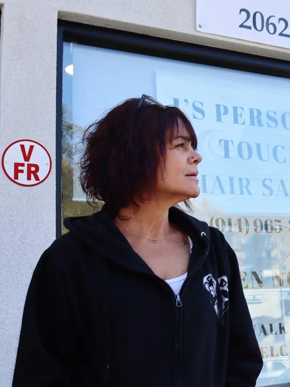 A woman with short, dark red hair standing outdoors near a window with a sign, wearing a black hoodie and sunglasses on her head.
