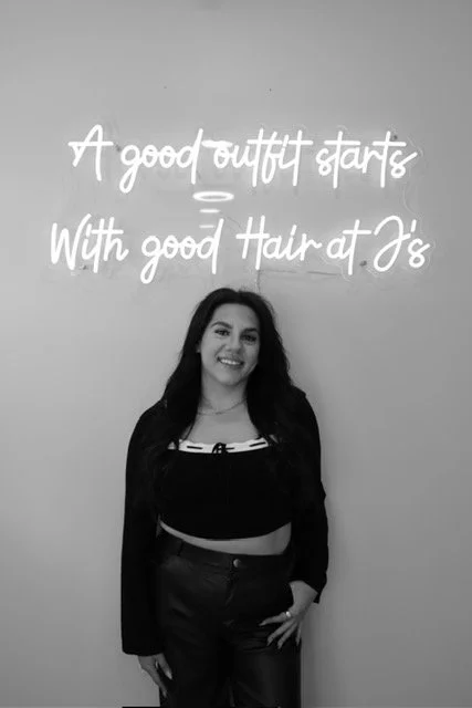 A young woman with long dark hair is standing in front of a light gray wall with white cursive writing that reads 'A good outfit starts with good hair and a smile'. She is wearing a black long-sleeve crop top, black leather pants, and is smiling with her hand on her hip.