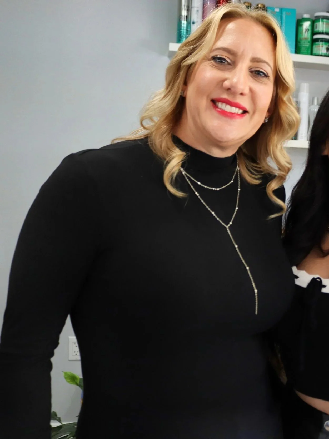 A woman with blonde hair smiling, wearing a black turtleneck and layered silver necklaces, standing in front of shelves with hair and beauty products.