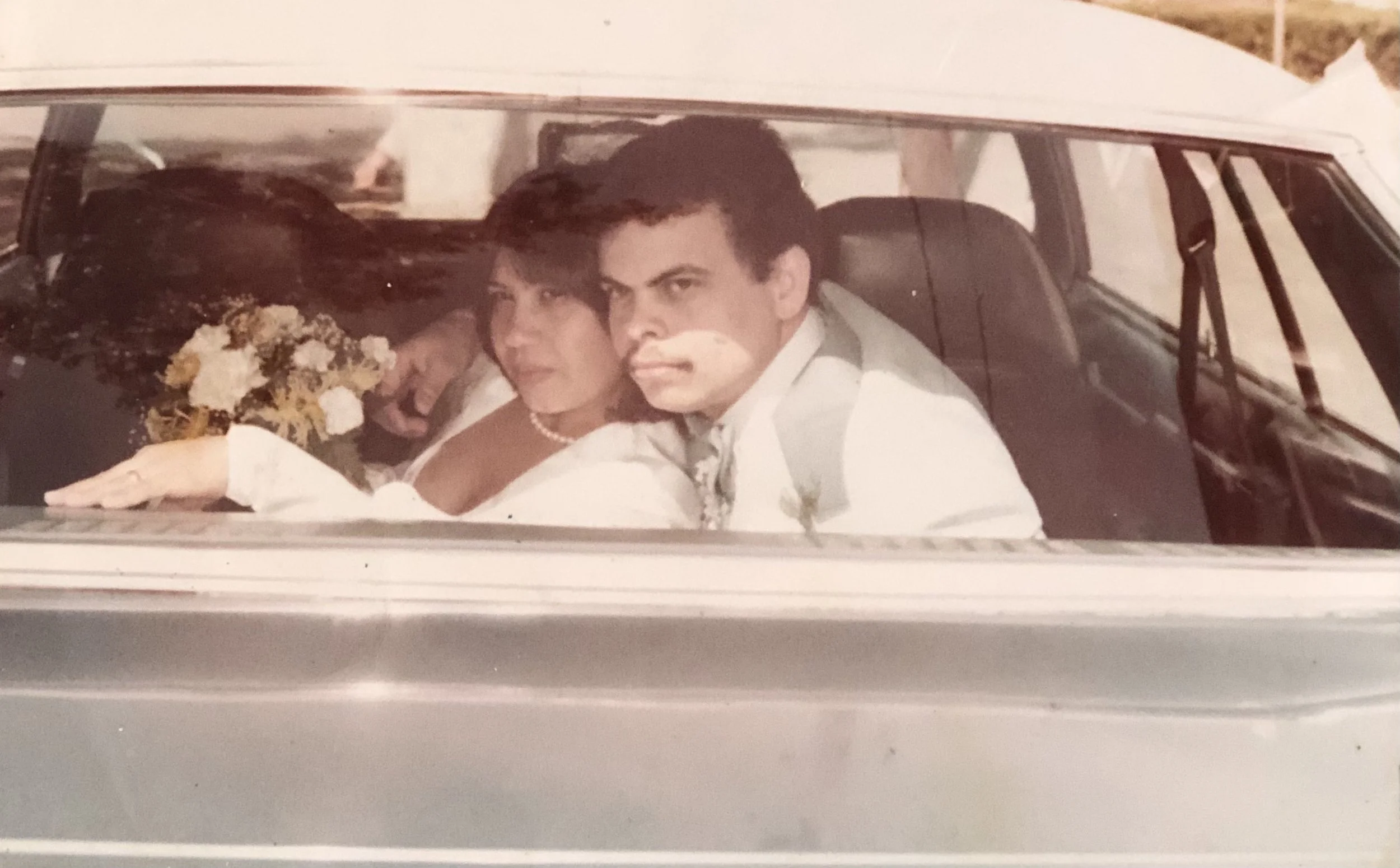 A black-and-white photo of a man and a woman sitting in the front seats of a car, with the woman holding a bouquet of flowers.