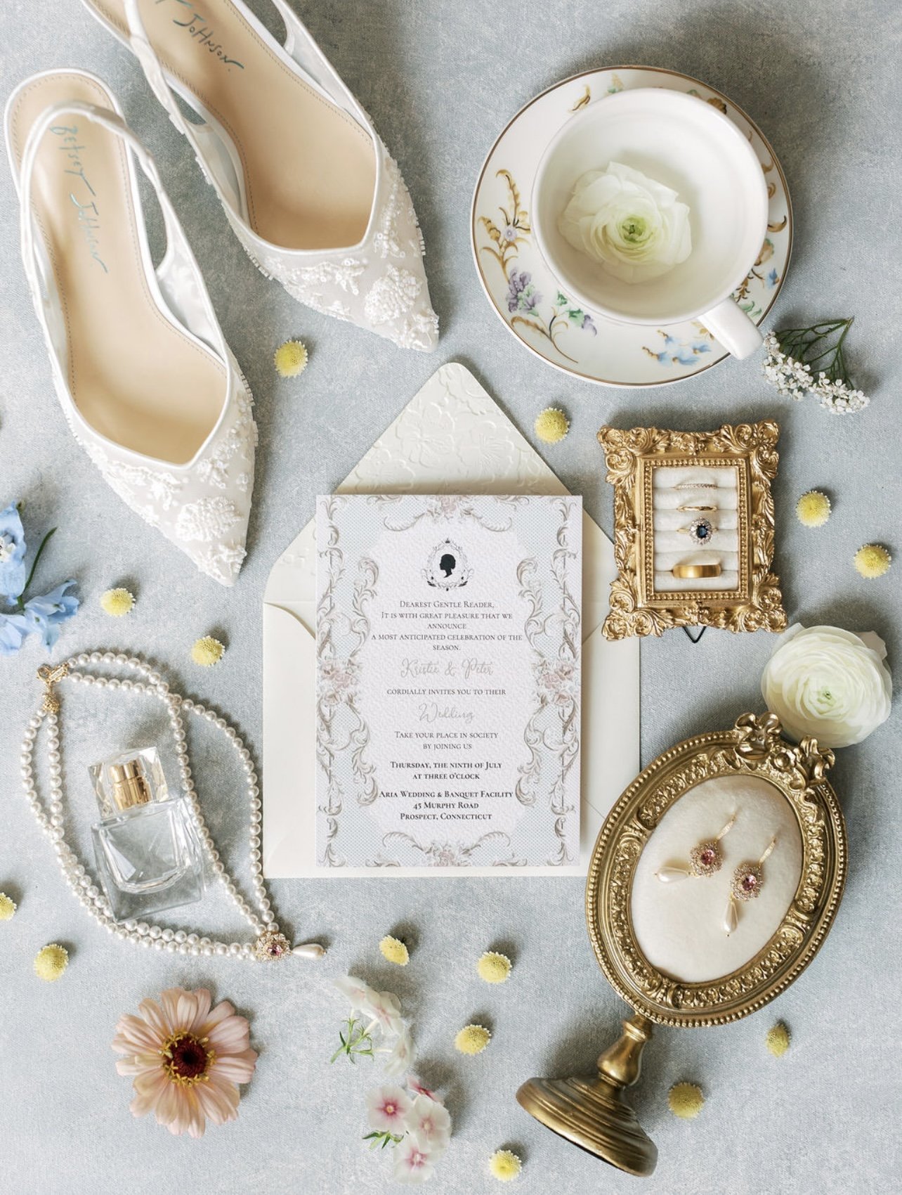 A wedding invitation surrounded by a bouquet of pink and purple flowers, a jewelry box with rings, a small bottle of Valentino perfume, and other decorative items on a lace-covered table.