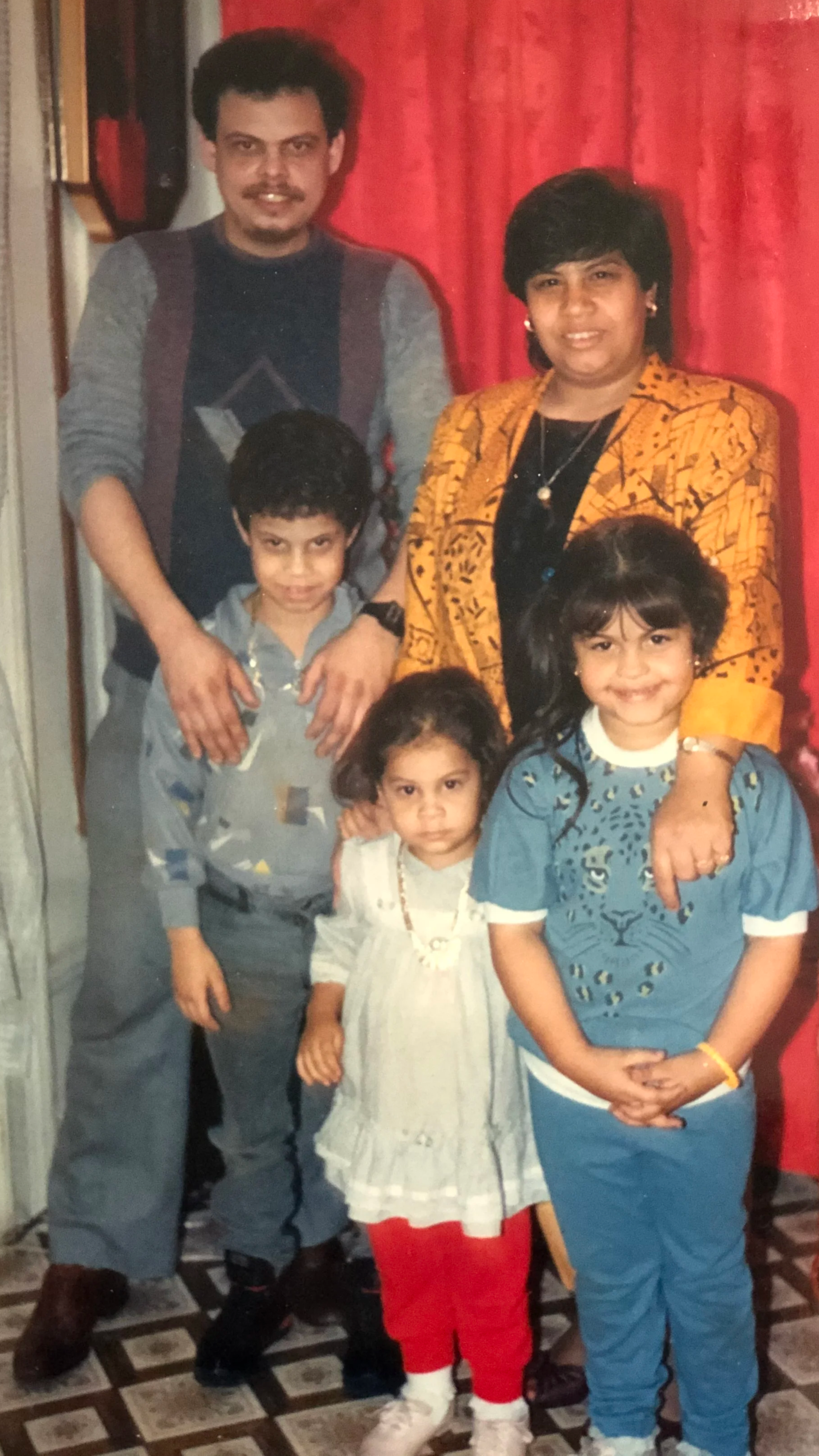 A family of six posing indoors in front of a red curtain. An adult man and woman, and four children, two girls and two boys, all smiling or with neutral expressions.