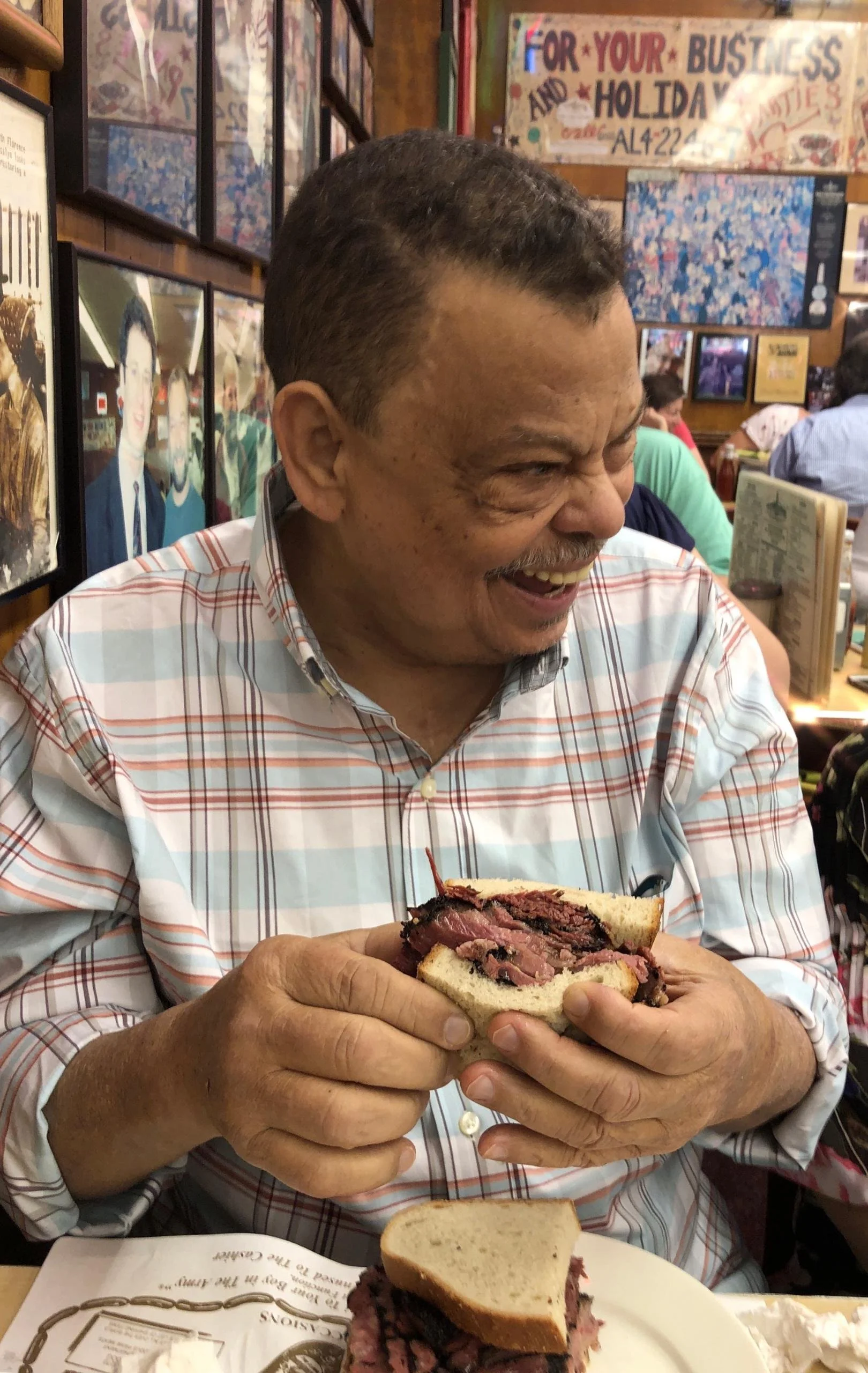 Man smiling and holding a large sandwich filled with sliced steak, in a lively restaurant with framed photos on the wall.