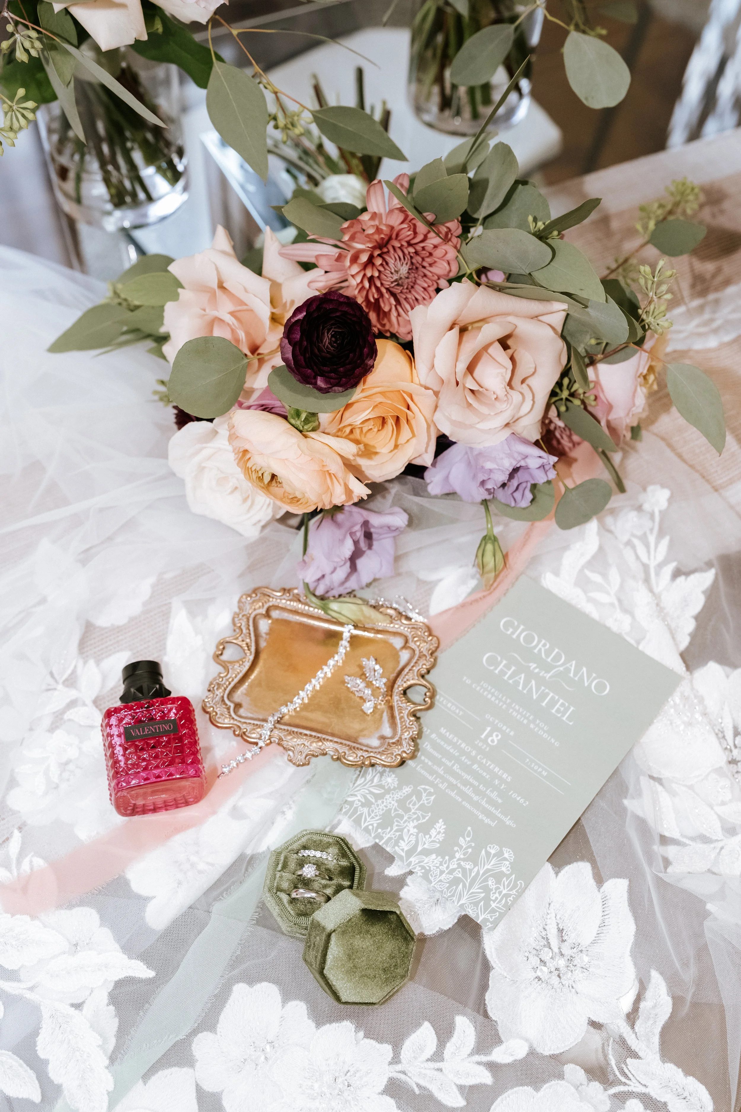 A wedding invitation surrounded by a bouquet of pink and purple flowers, a jewelry box with rings, a small bottle of Valentino perfume, and other decorative items on a lace-covered table.