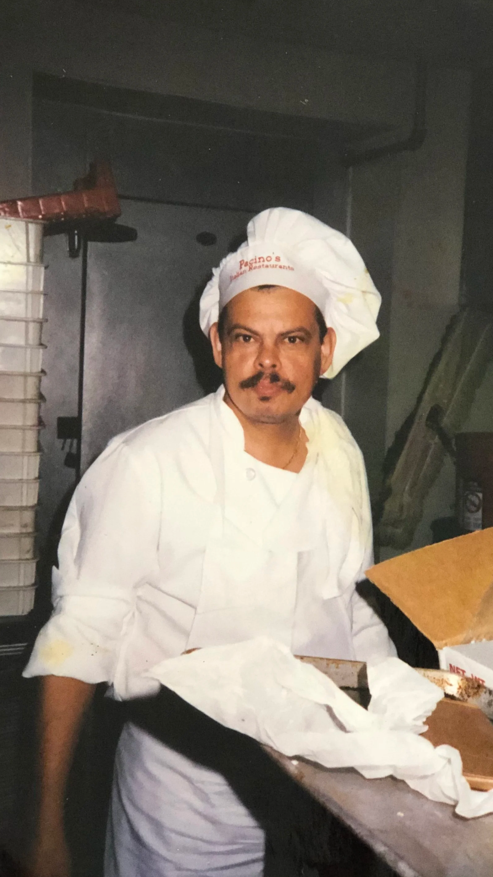 A male chef with a mustache and beard wearing a white chef's uniform and a chef hat, standing in a kitchen with various kitchen utensils and appliances in the background.
