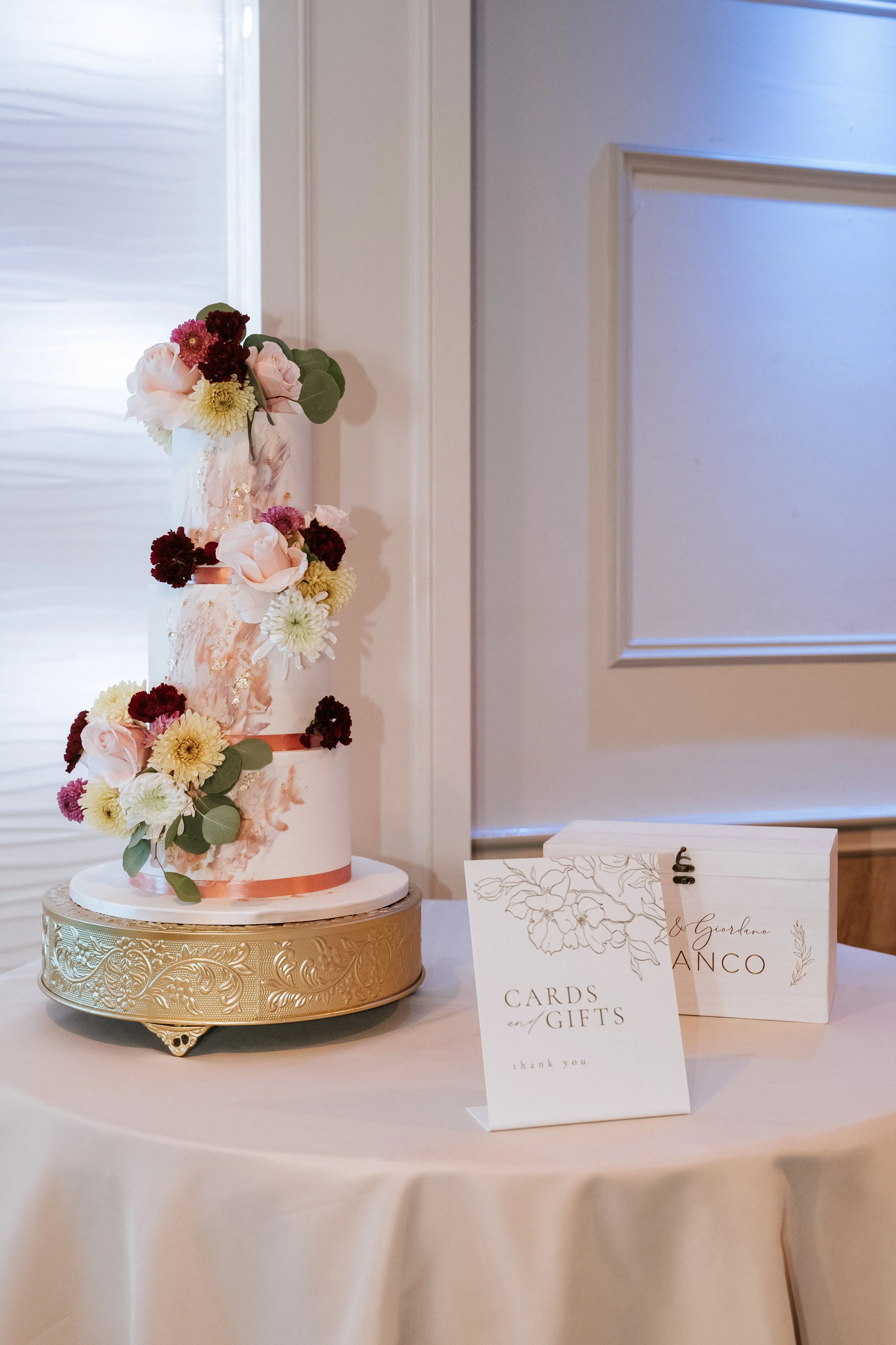 A three-tiered wedding cake decorated with pink and white flowers, including roses and chrysanthemums, with a gold cake stand on a beige tablecloth. There are two white signs, one reading 'CARDS AND GIFTS' and the other with a name, placed on the table.