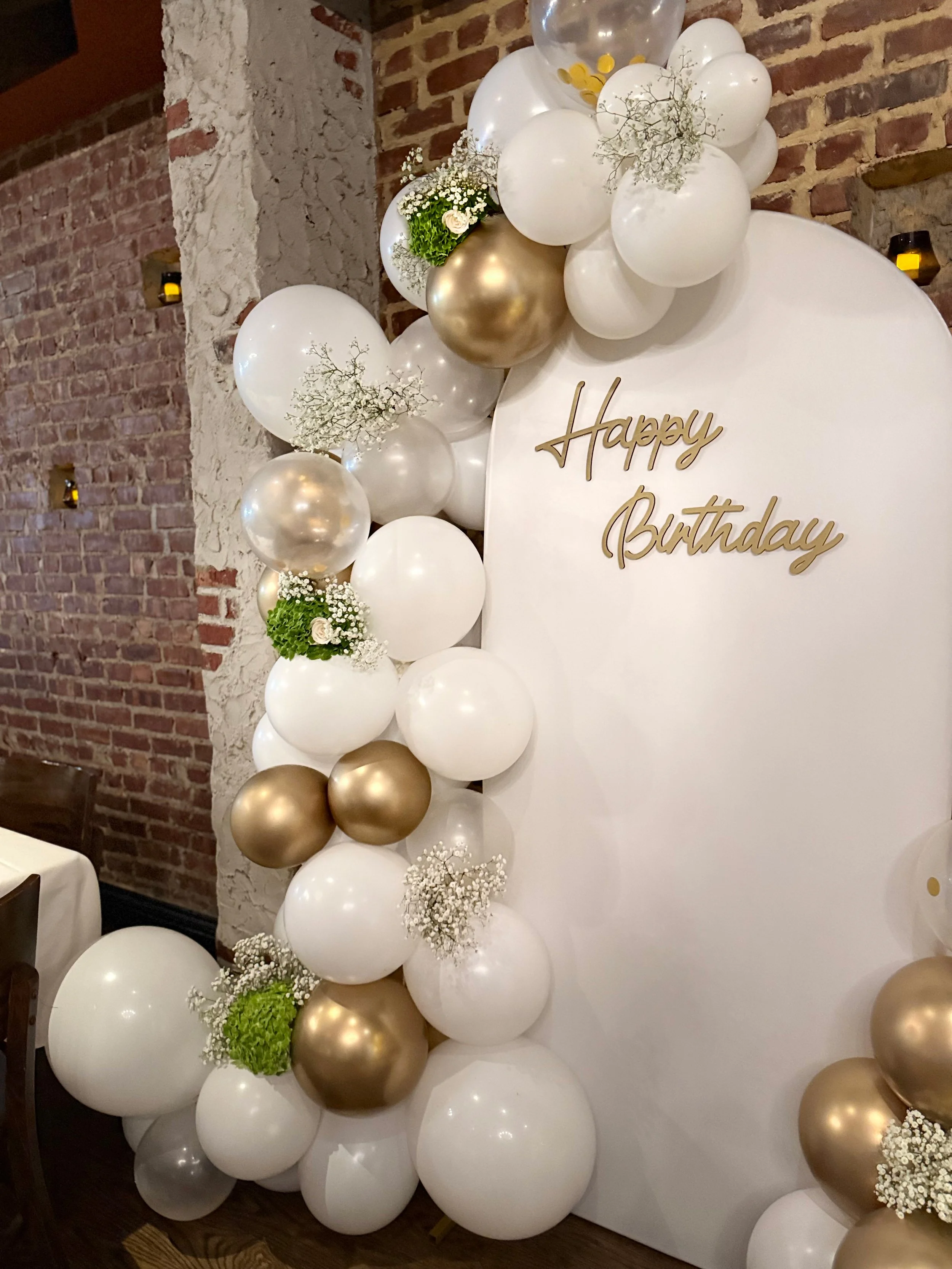 Decorative balloon arrangement with white, gold, and clear balloons, small white flowers, green foliage, and a gold "Happy Birthday" sign on a white backdrop.